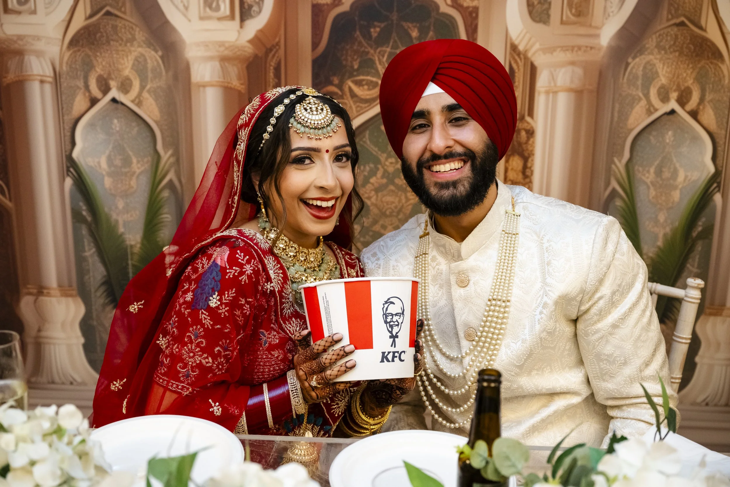 A bride and groom at their wedding celebration, smiling and holding a KFC bucket, with traditional attire and jewelry in a decorated wedding venue.