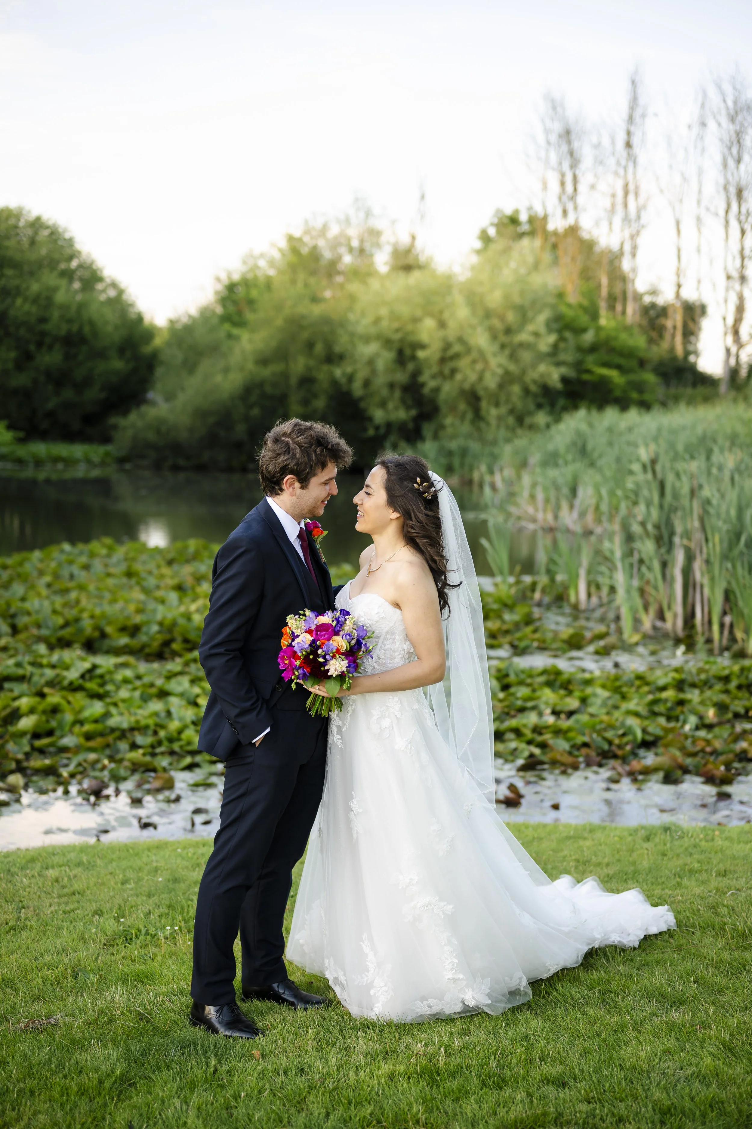 A bride and groom standing facing each other outdoors by a pond, holding a bouquet of colorful flowers, with a background of trees and water lilies.