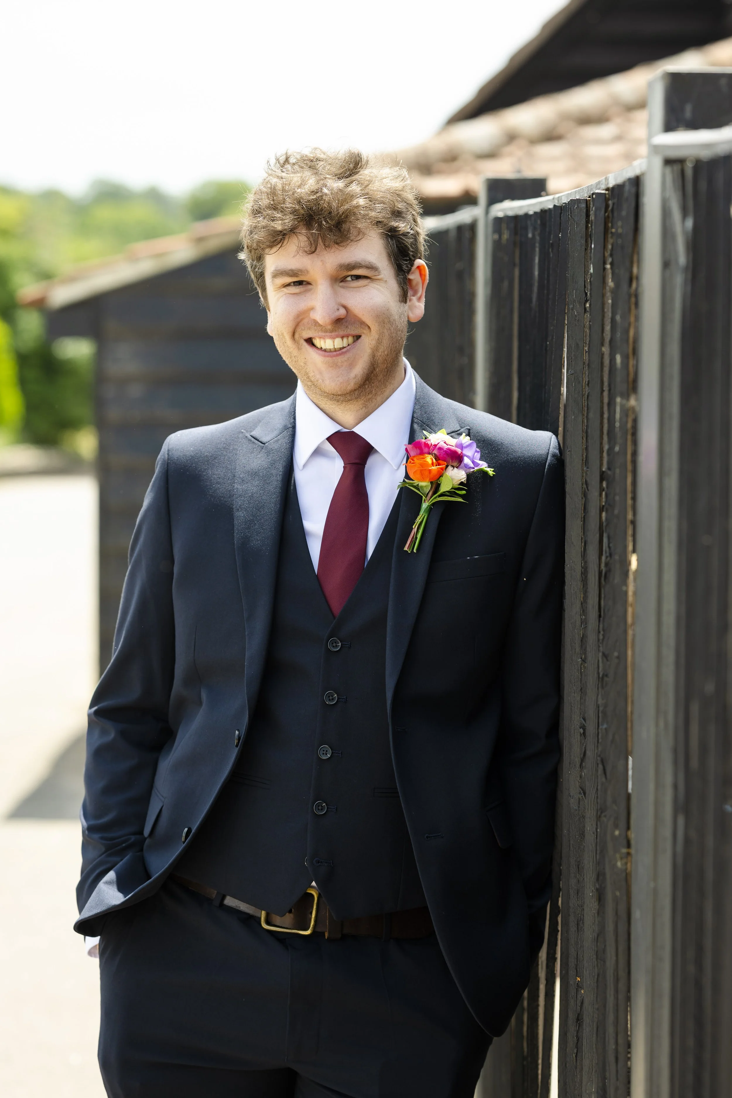 A young man in a black suit with a burgundy tie and a colorful boutonniere, smiling and leaning against a wooden fence outdoors.