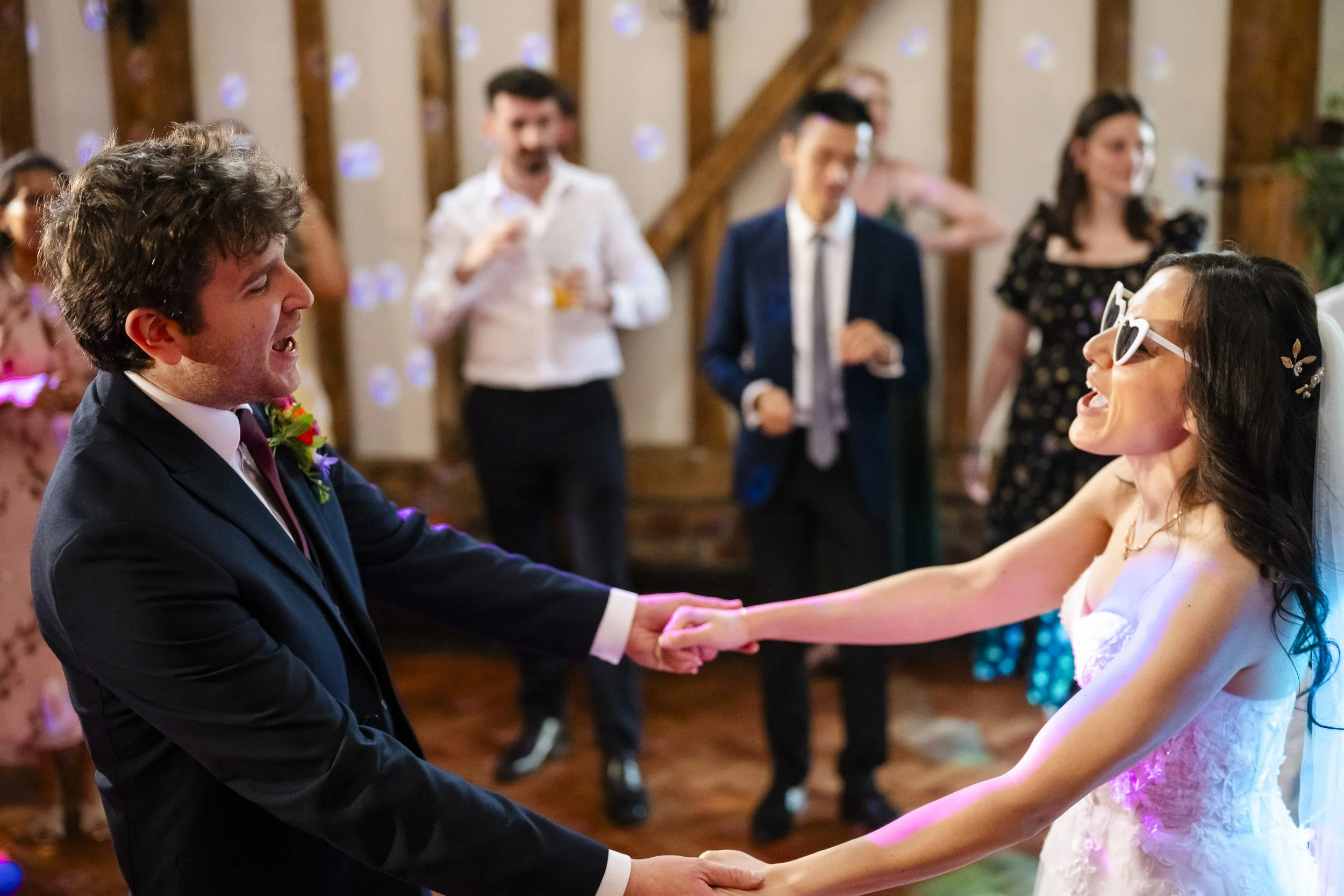 A bride and groom dancing holding hands at their wedding reception, surrounded by guests.