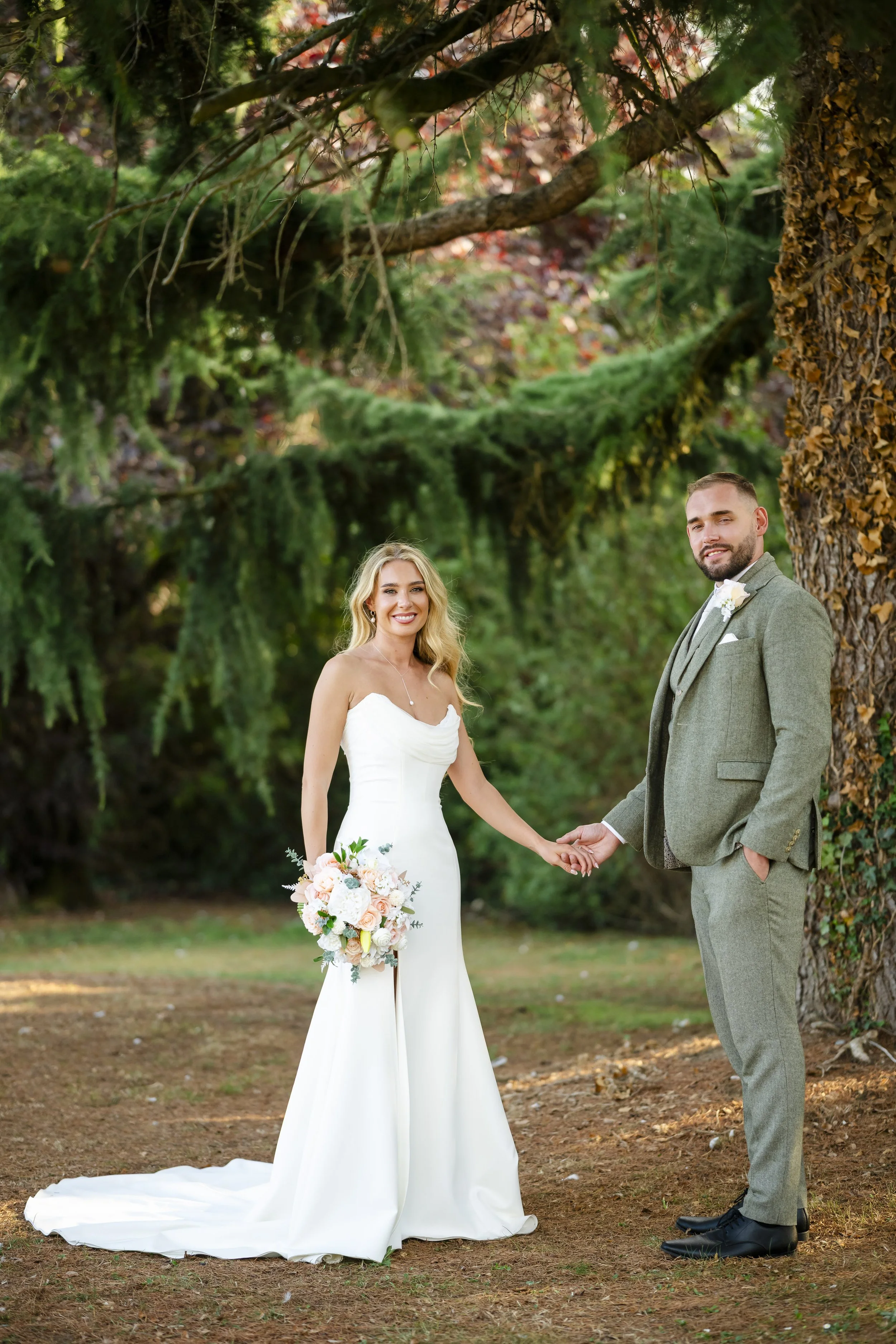 A newlywed couple holding hands outdoors in a forest setting, the woman in a white wedding gown and holding a bouquet, and the man in a gray suit with a boutonniere.