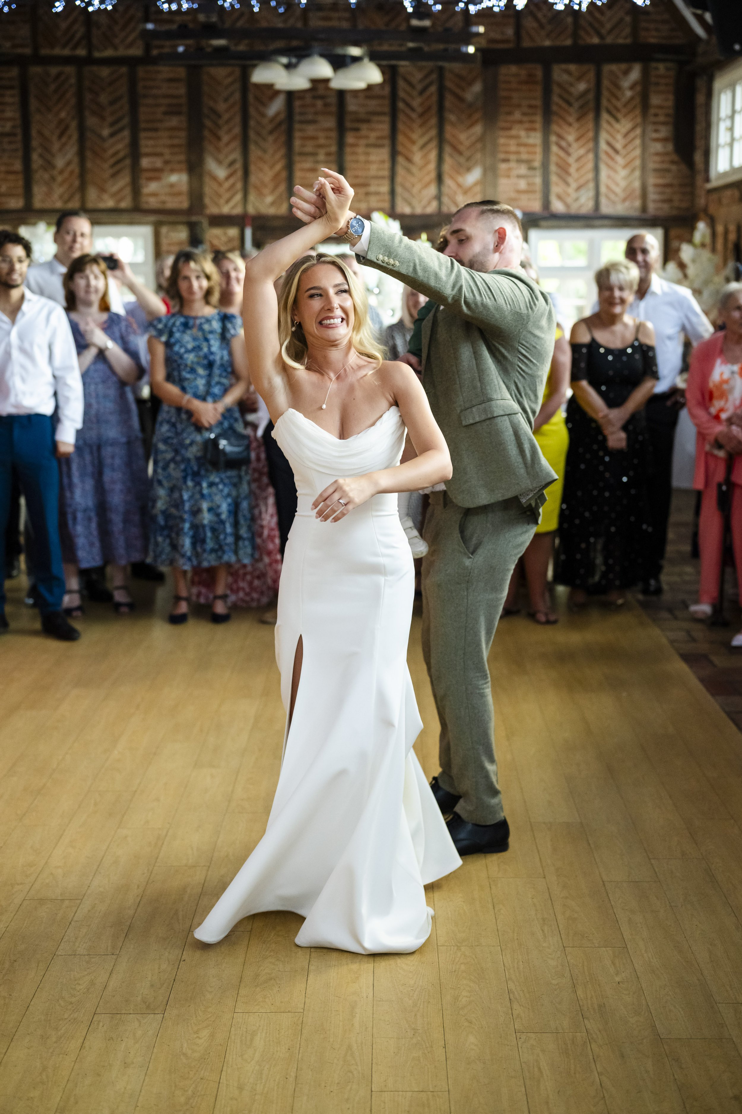 A bride and groom are dancing at their wedding reception, surrounded by family and friends in a rustic indoor venue with wood walls and high ceilings.