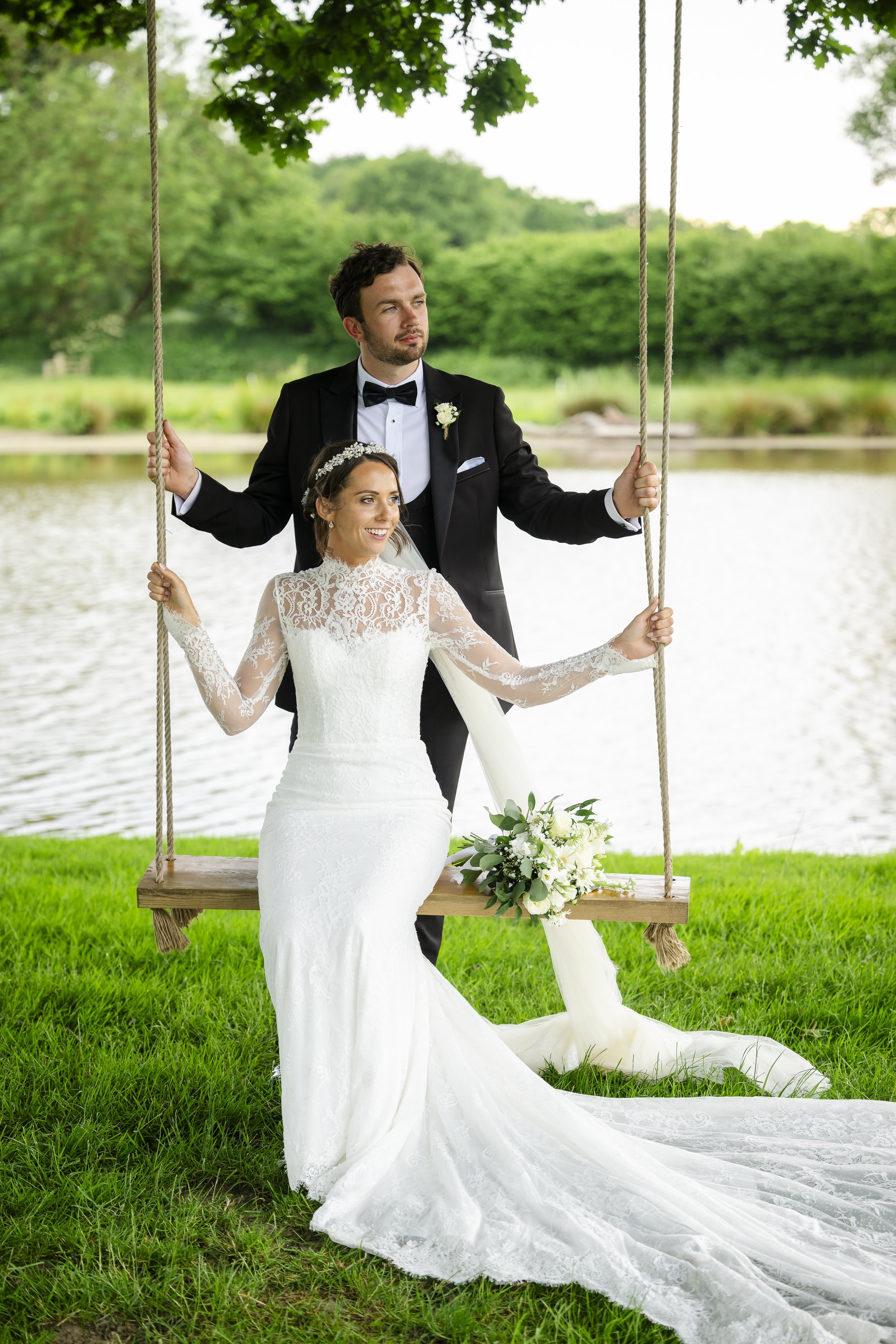 A bride in a white lace wedding dress and a groom in a tuxedo stand on a grassy area by a lake, holding onto the ropes of a wooden swing.