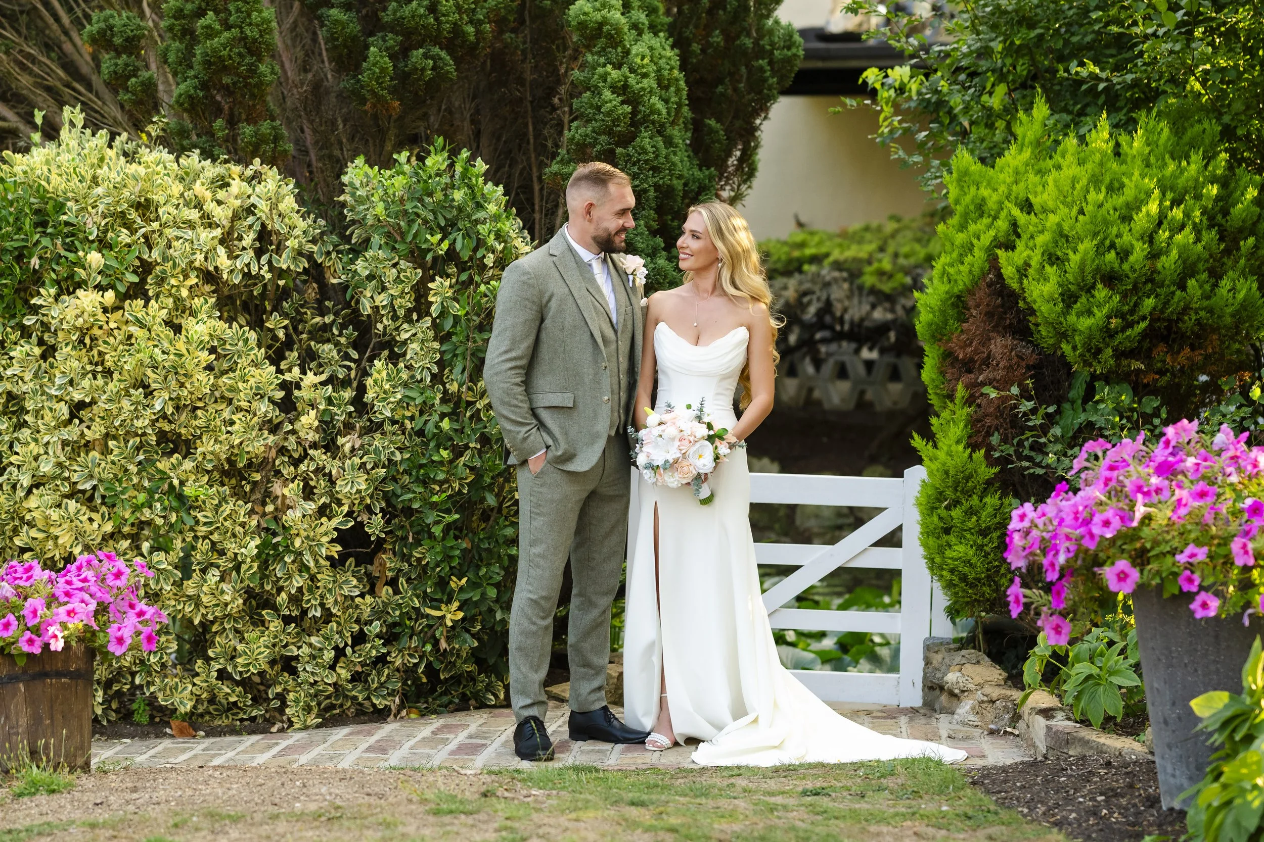 A bride and groom stand together outdoors, surrounded by lush green bushes and vibrant pink flowers. The bride wears a strapless white wedding gown with a slit, holding a bouquet of white and pink flowers. The groom is dressed in a light gray suit wi