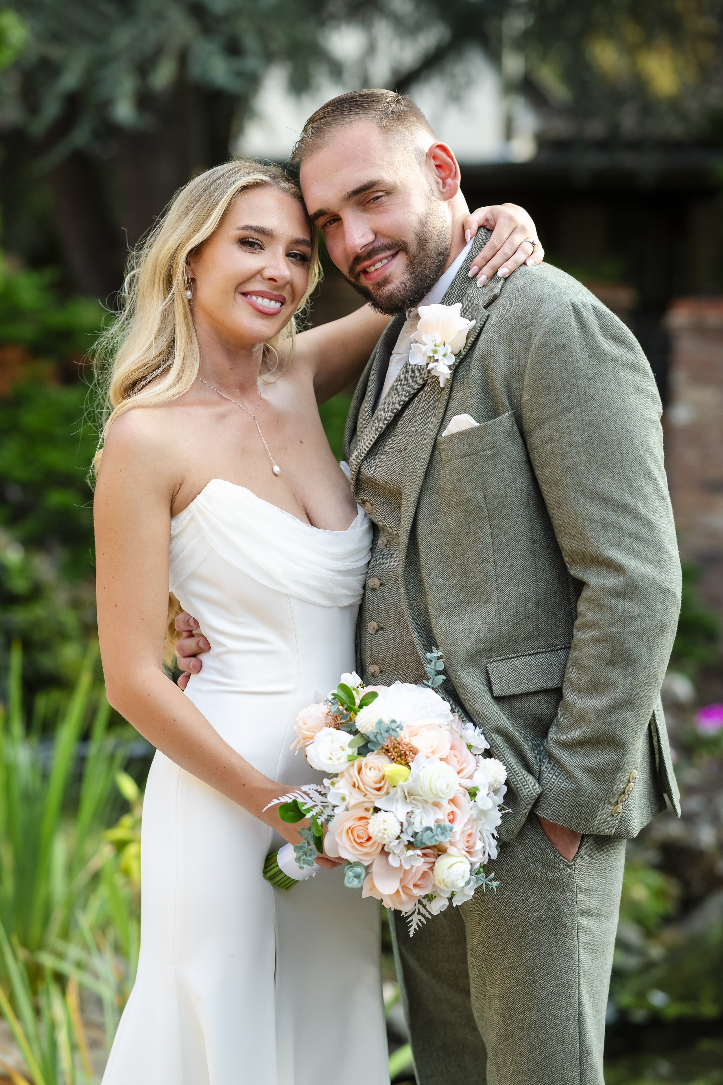 A bride and groom posing outdoors in wedding attire, with the bride holding a bouquet of flowers and the bride resting her hand on the groom's neck.
