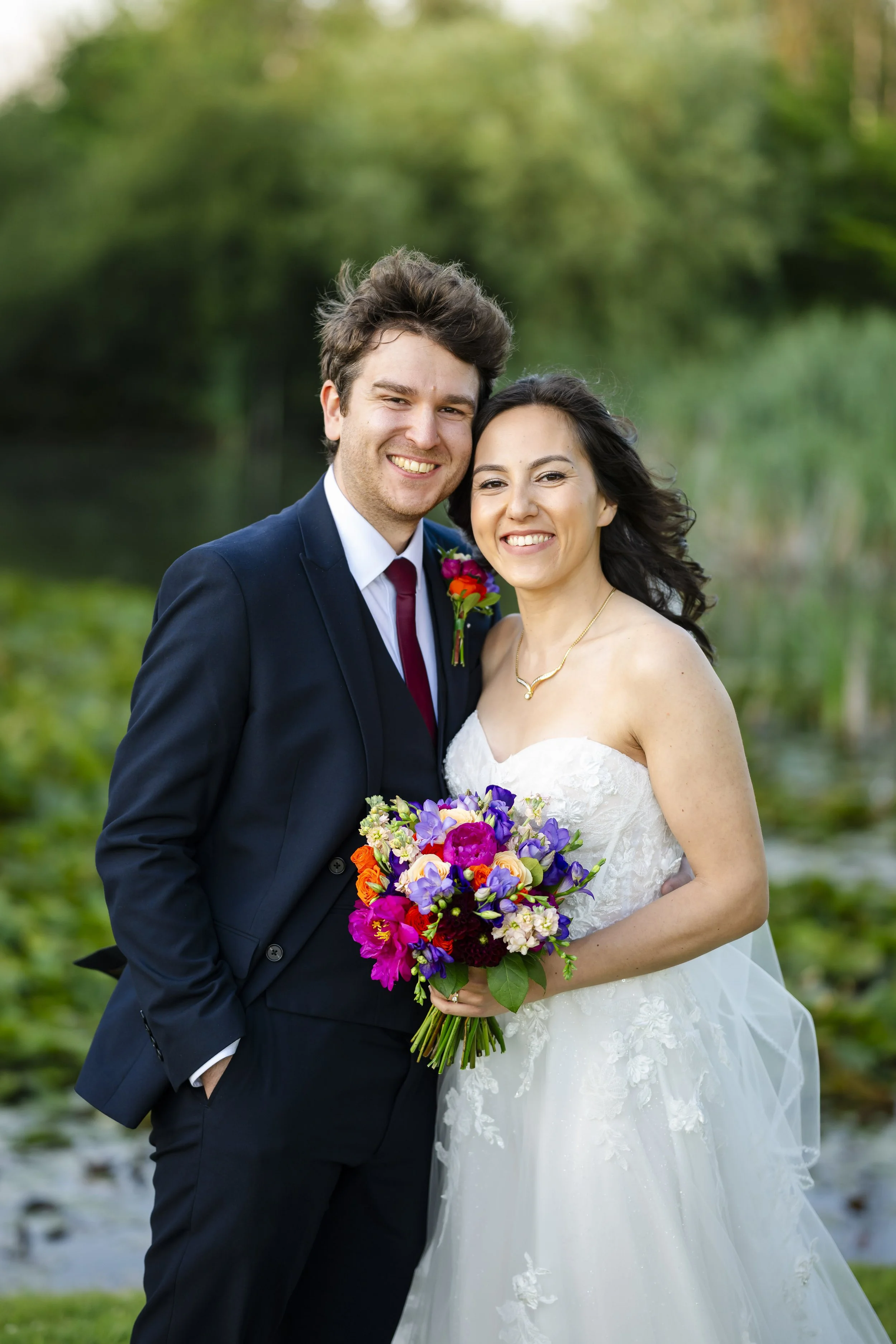 A newlywed couple smiling outdoors, with the groom in a navy suit and the bride in a white wedding dress holding a colorful bouquet.