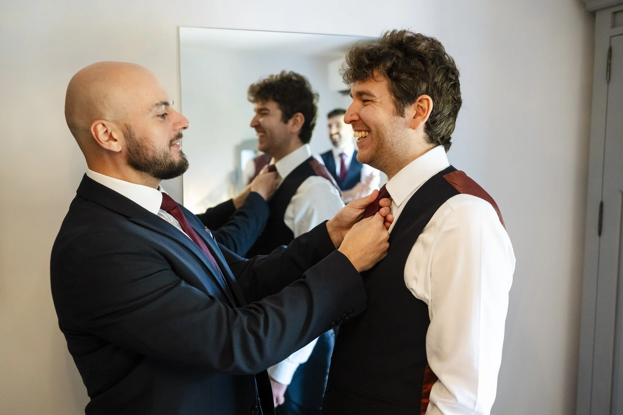Two men in suits dressing a third man in a vest and shirt, with a mirror reflecting their smiles.