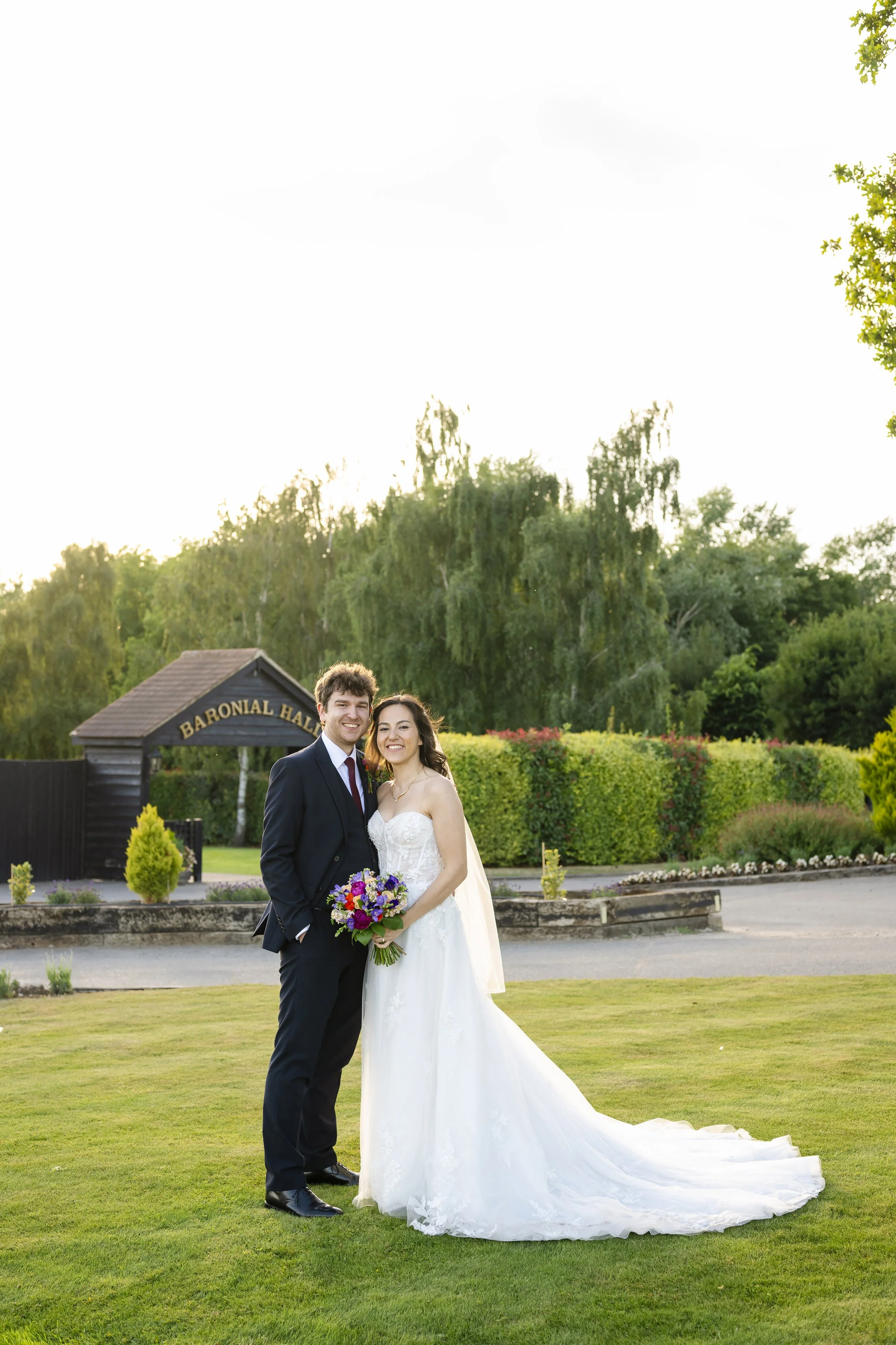 A newlywed couple standing on a grassy area outside at sunset, with a wedding bouquet, in front of a sign that reads 'Baronial Hall,' surrounded by trees and bushes.