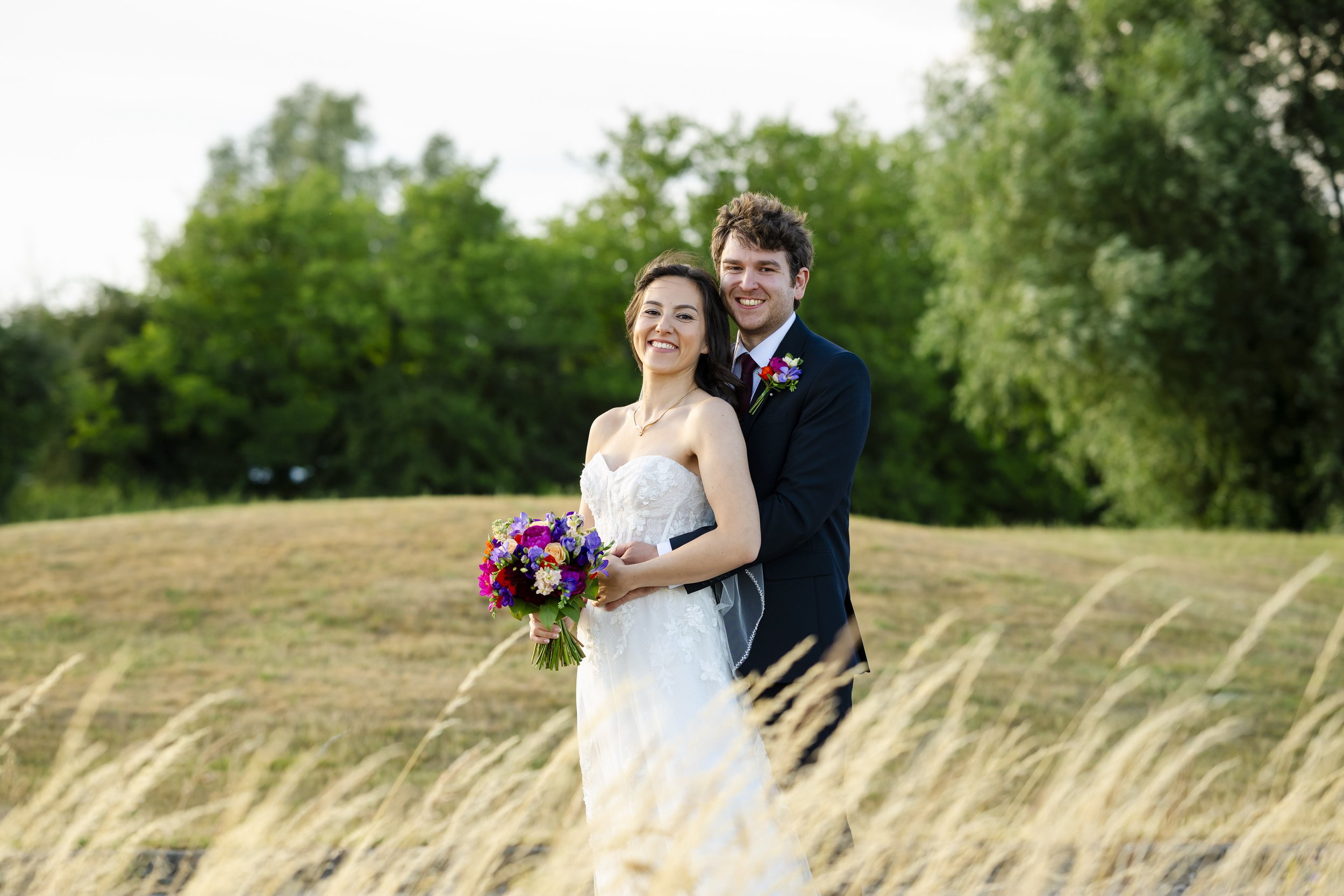 A happy bride and groom in wedding attire standing outdoors in a grassy field with green trees in the background. The bride is holding a colorful bouquet and the groom has a boutonniere. They are smiling at the camera.