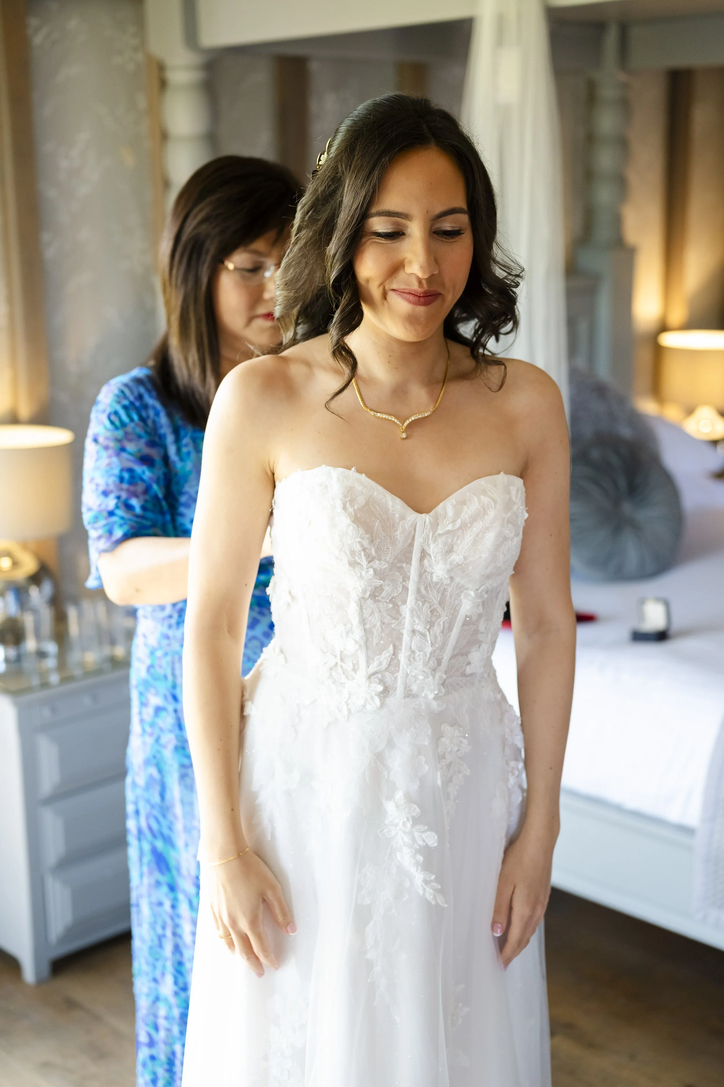 A bride in a white strapless wedding gown standing in a bedroom, being helped with her dress by another woman in a blue dress.