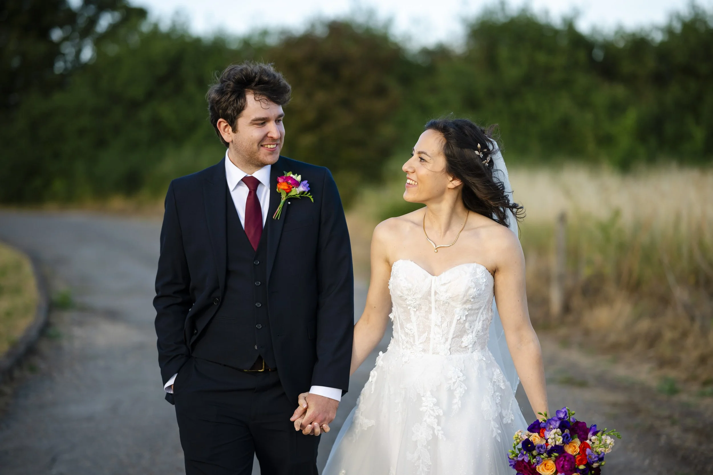 A newlywed couple walking outdoors, smiling at each other, holding hands. The bride is in a white lace gown holding a colorful bouquet, and the groom is dressed in a dark suit with a red tie and a flower boutonniere. There are trees in the background