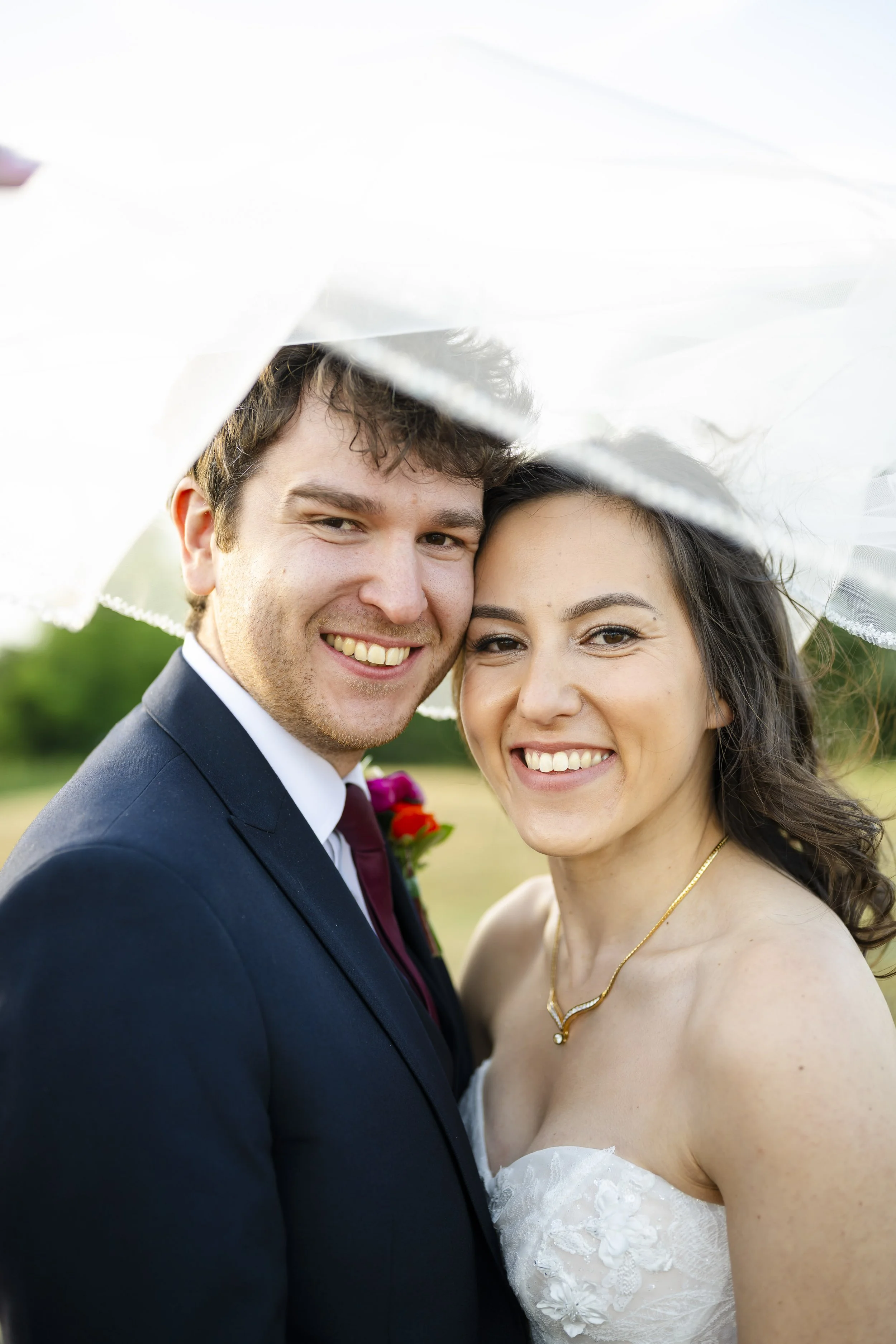 A smiling bride and groom under a veil outdoors on a wedding day.