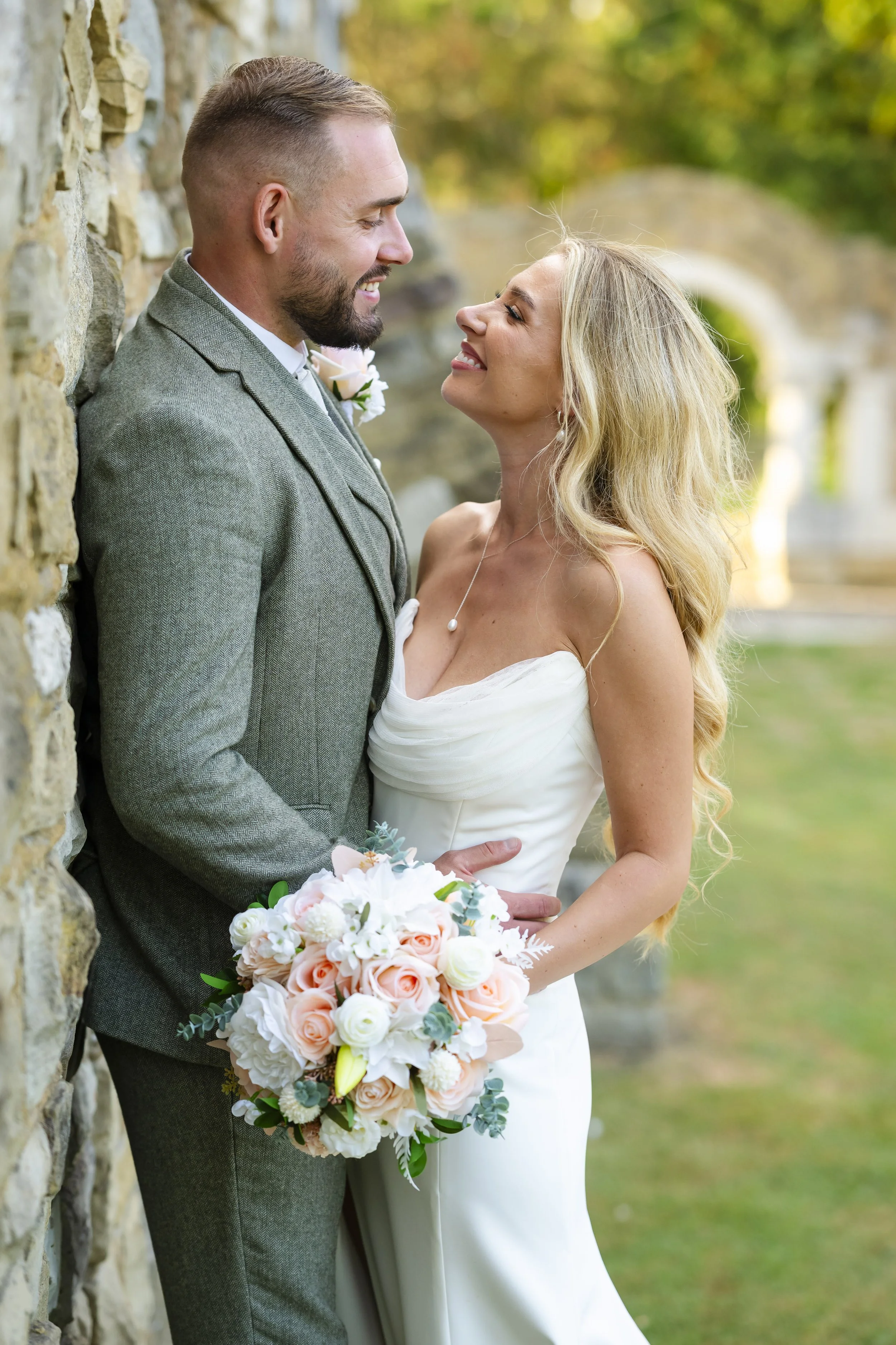 A newlywed couple faces each other, smiling, outdoors, with a stone wall and lush greenery in the background. The bride holds a bouquet of pink and white roses with greenery.