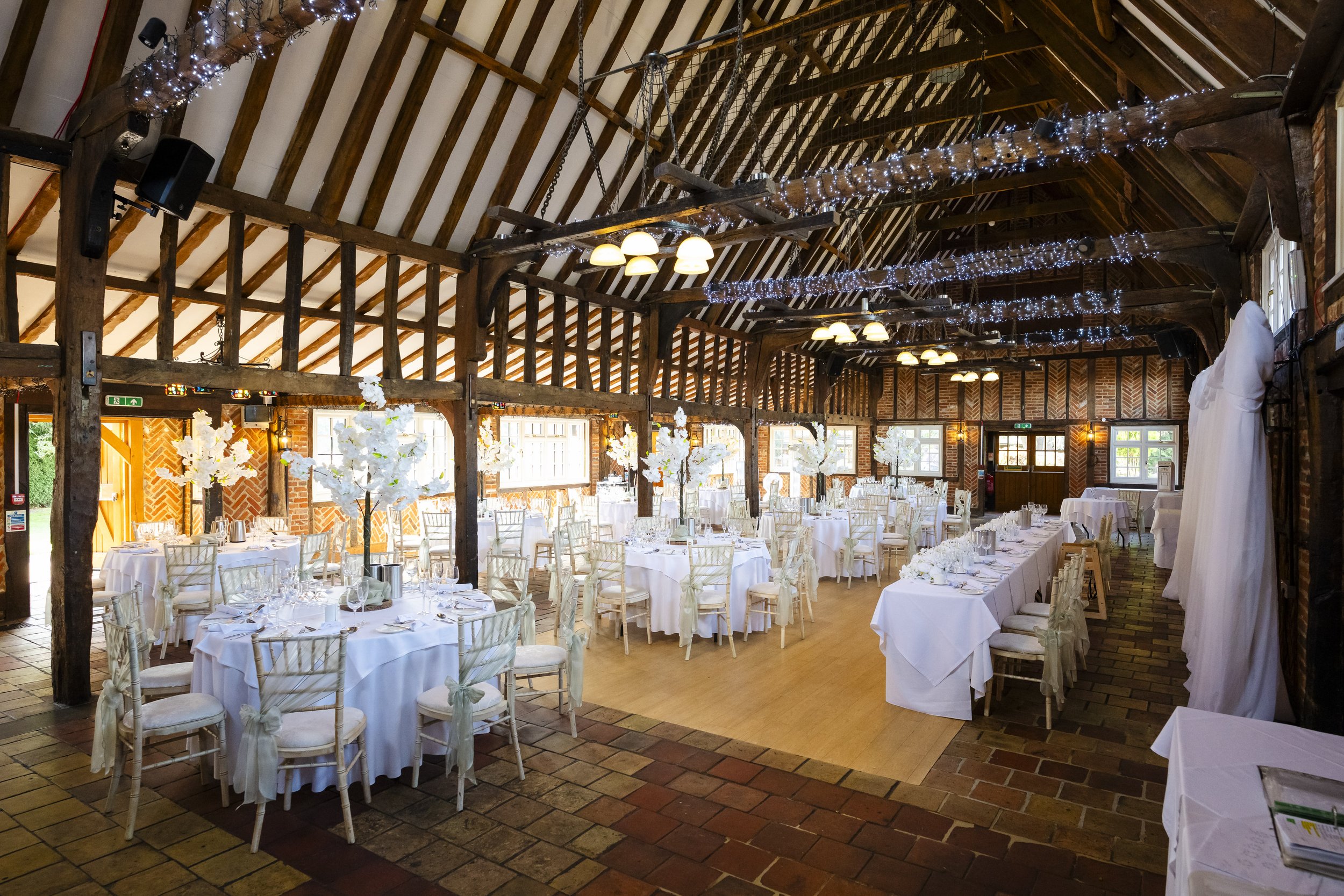 Wedding reception setup inside a rustic barn with wooden beams, decorated with fairy lights, white floral centerpieces, and elegantly arranged tables and chairs.