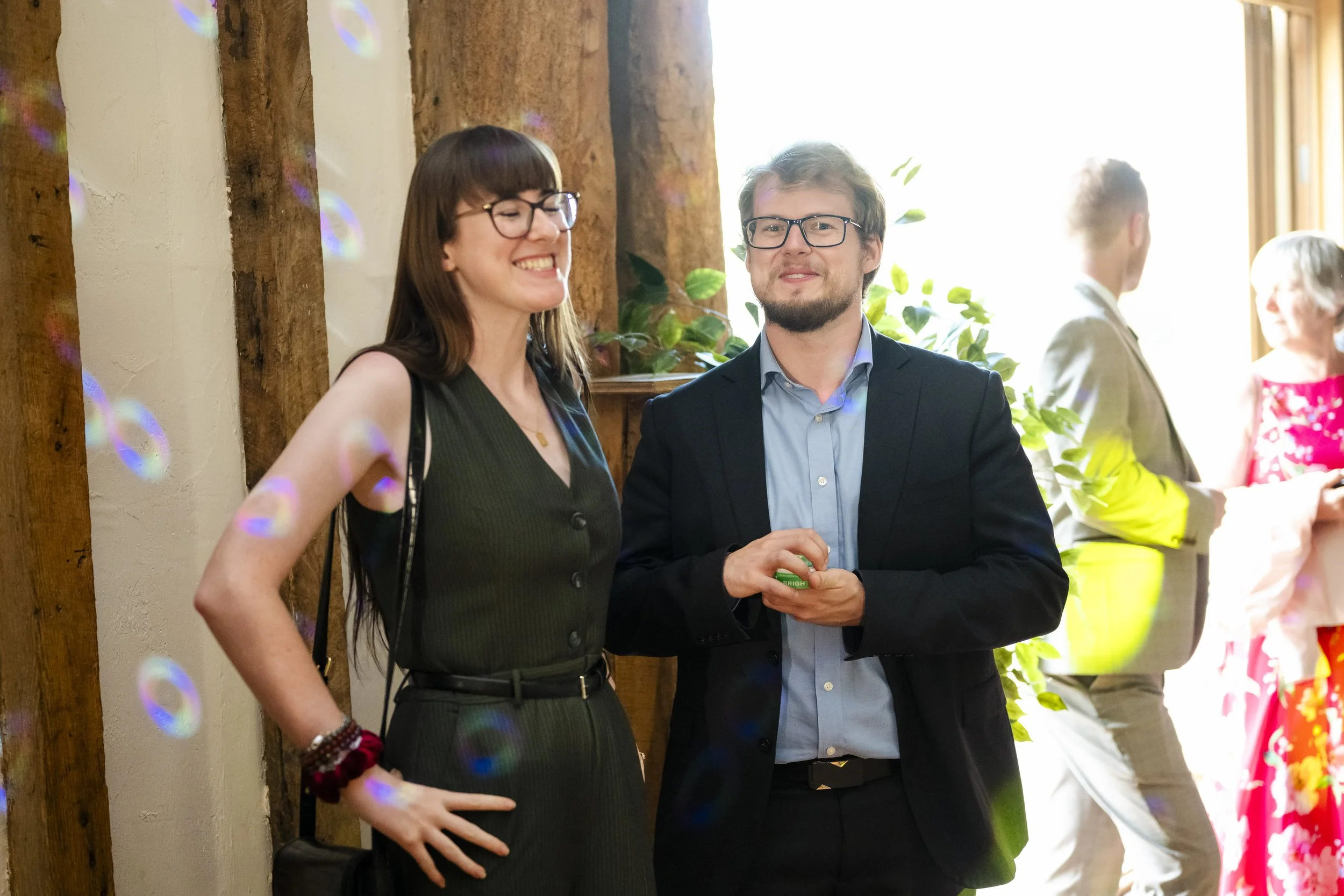 Two people, a woman with glasses and a man with glasses, talking and smiling at a social gathering, standing near a wooden wall with sunlight behind them.