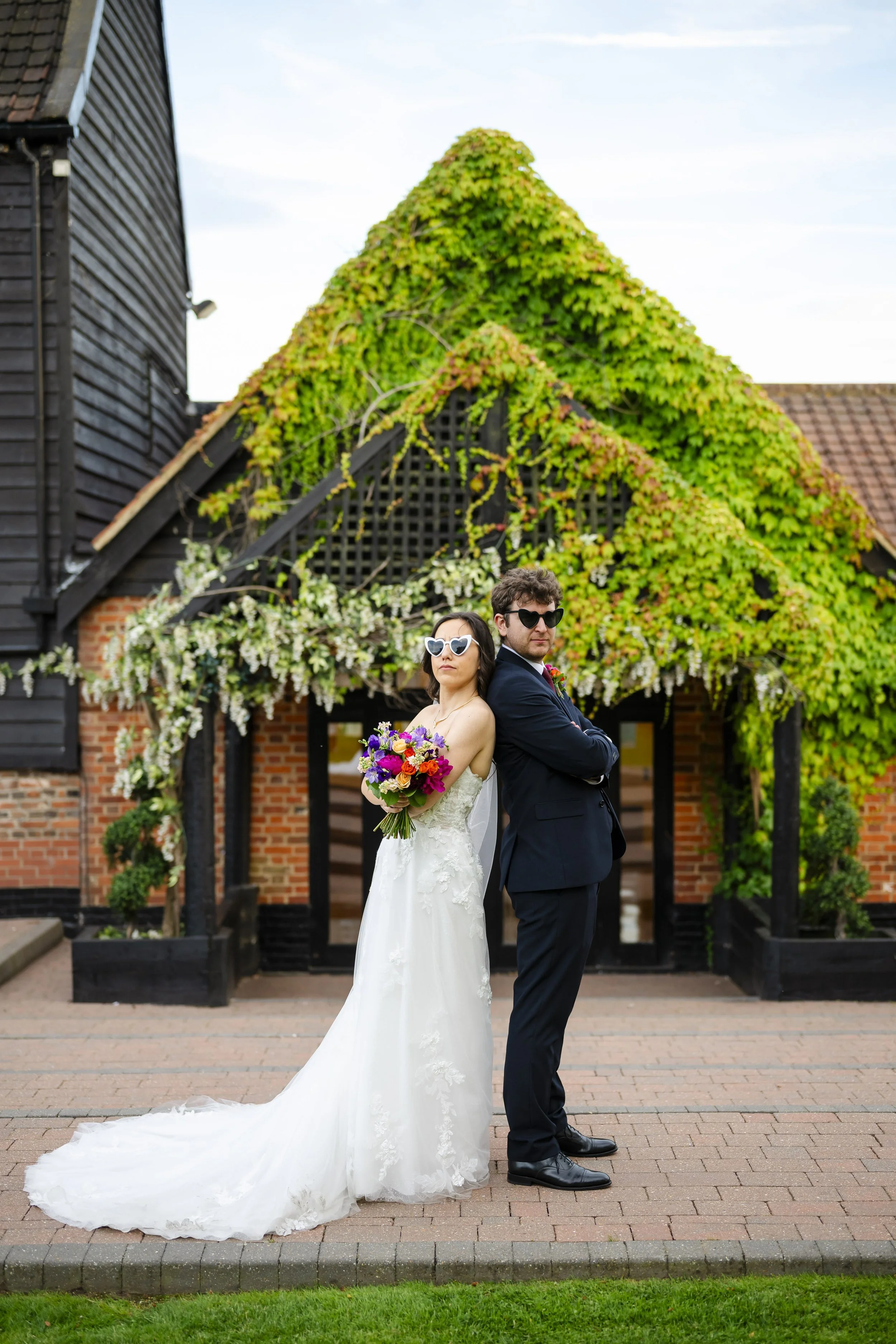 Bride and groom standing back-to-back in front of a house with green vines and flowers, both wearing sunglasses; bride holding a colorful bouquet, groom crossing his arms.
