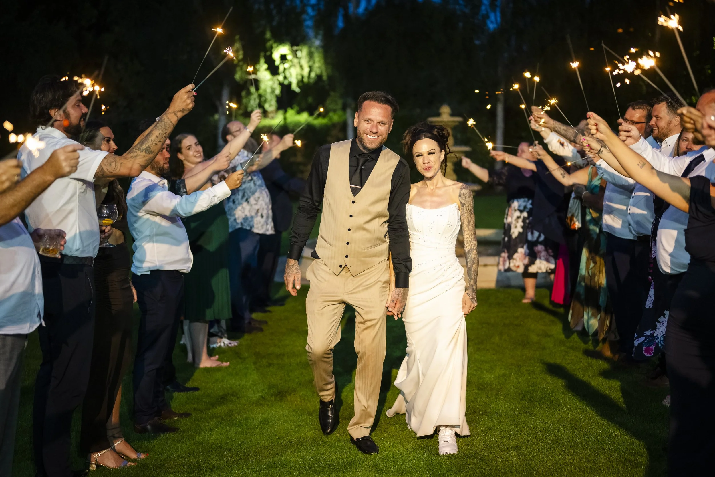 A newlywed couple walking hand in hand under a night sky, surrounded by friends and family holding sparklers at a wedding celebration.