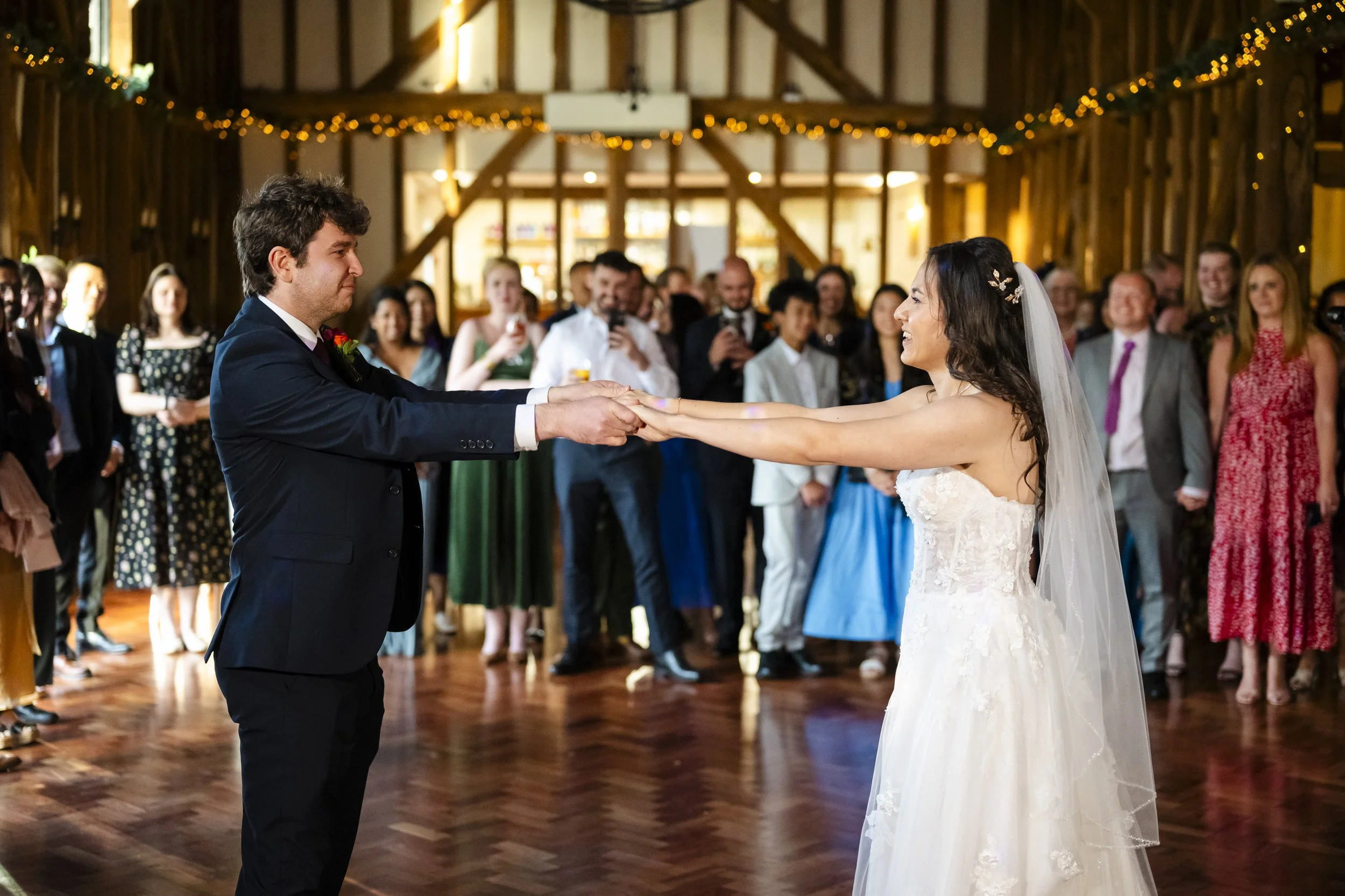 A bride and groom holding hands and smiling during their wedding dance in a barn decorated with string lights while guests look on warmly.