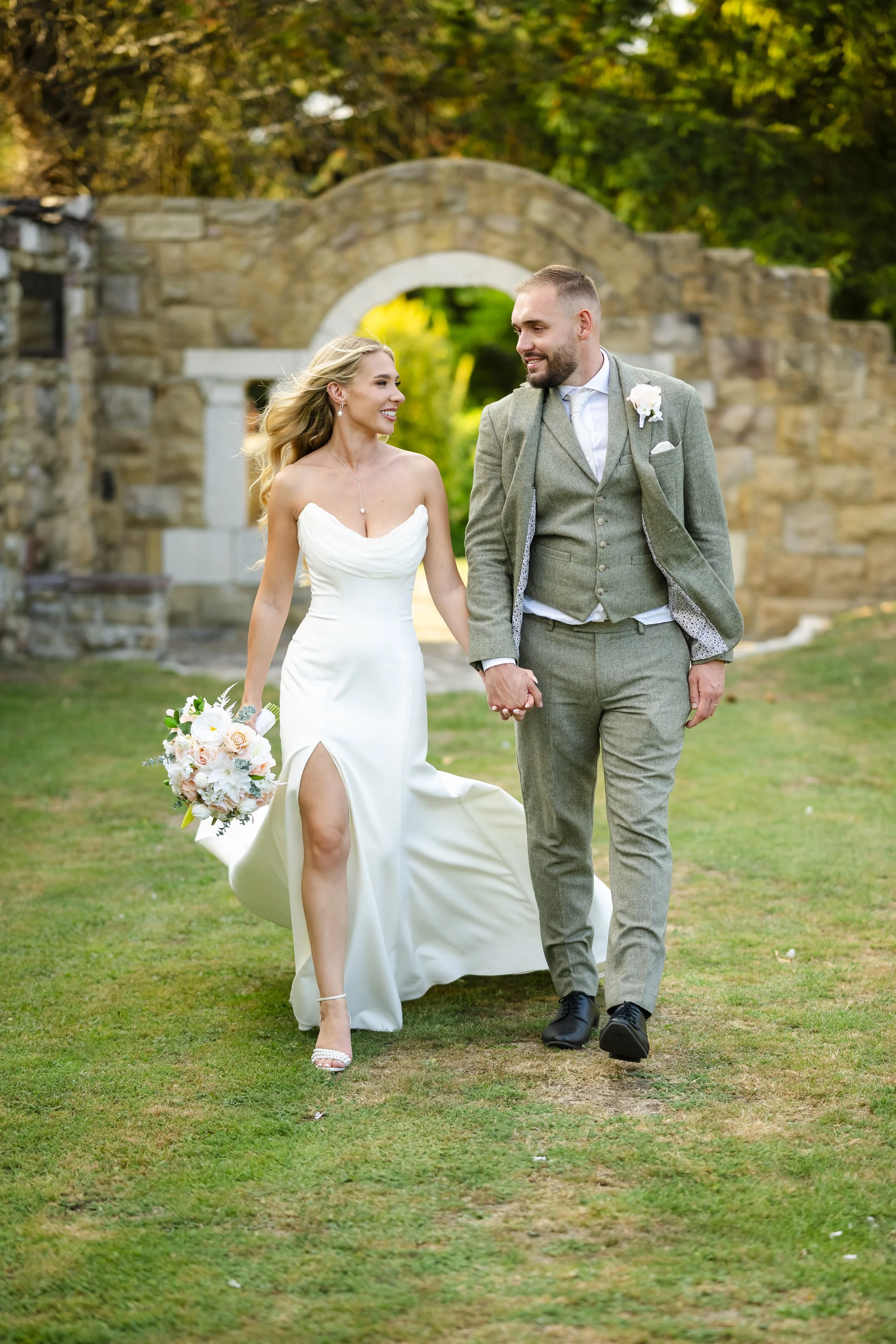 A bride and groom walking hand in hand outdoors, smiling and looking at each other, during their wedding. The bride has blonde hair and is wearing a white strapless wedding dress with a thigh-high slit, holding a bouquet of white and blush flowers. T