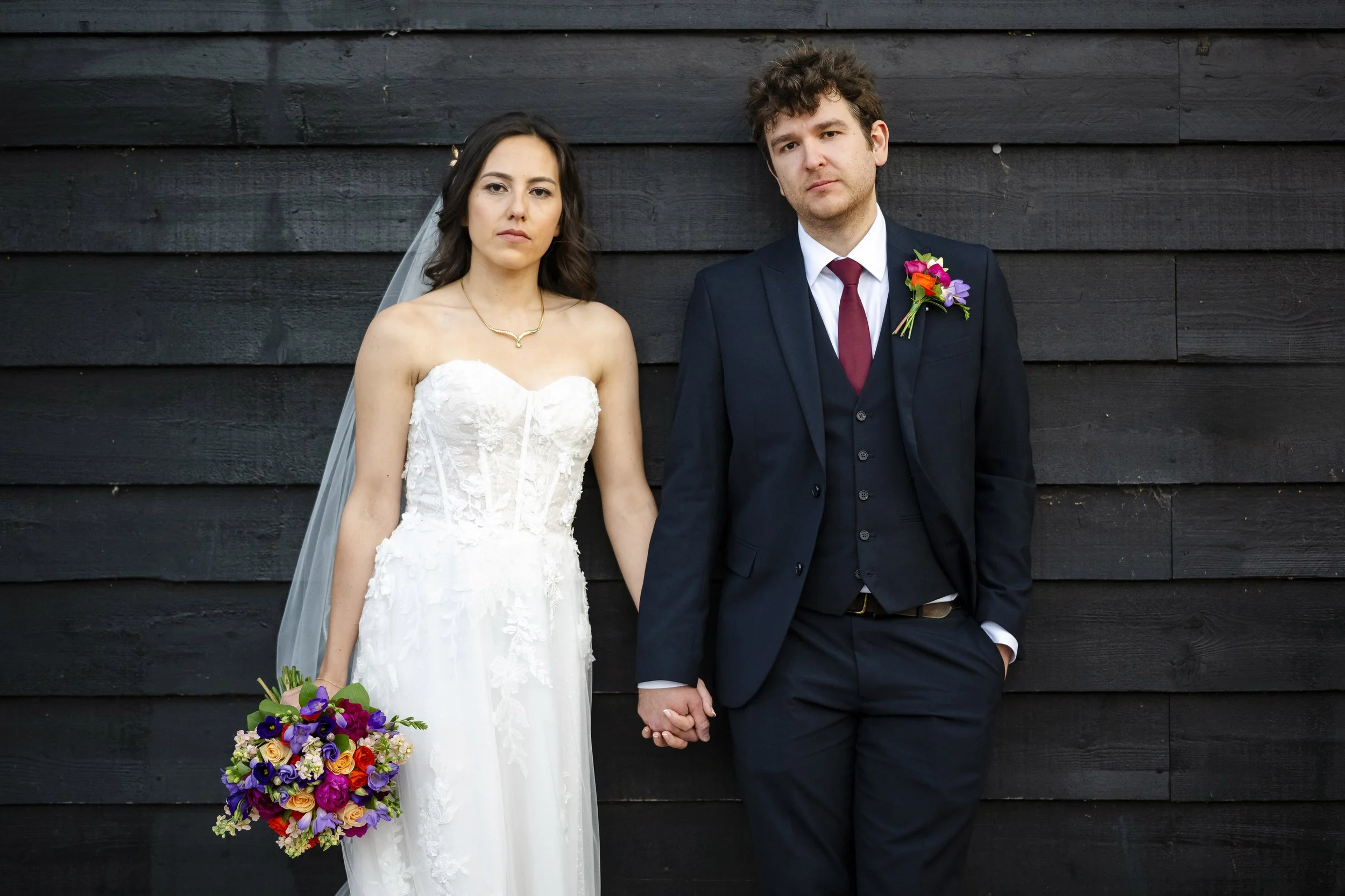 A bride and groom standing side by side holding hands, the bride in a white wedding dress with a floral bouquet, and the groom in a dark suit with a boutonniere, against a black wooden wall background.
