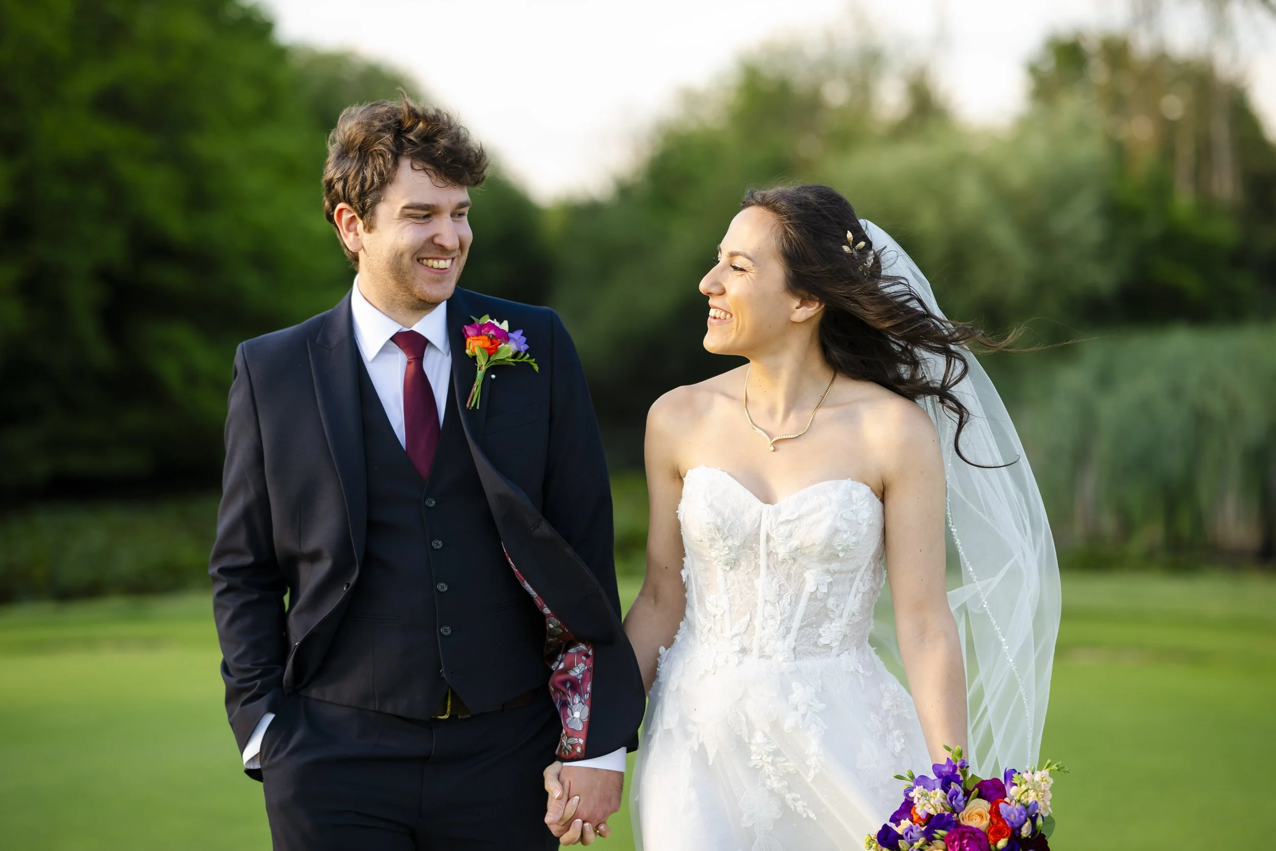 A bride and groom holding hands and smiling at each other outdoors during a wedding ceremony. The groom wears a dark suit with a red tie and colorful boutonniere. The bride wears a strapless white wedding dress, veil, and a purple floral bouquet.