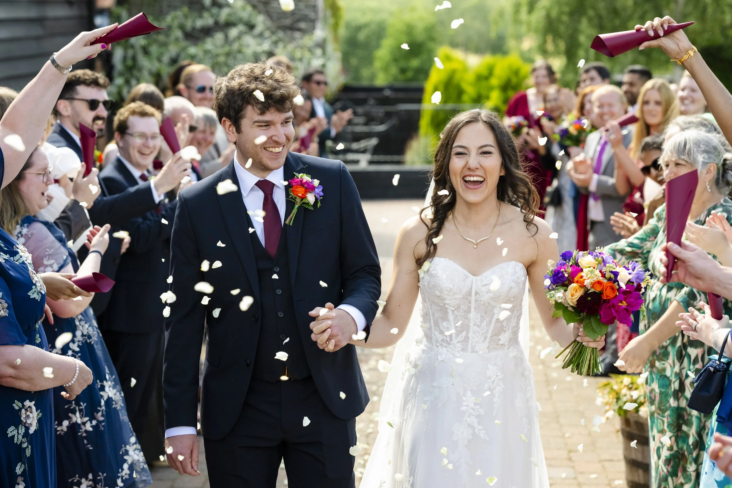 A newlywed couple walking hand in hand through a joyful crowd, throwing flower petals at a wedding celebration outdoors.