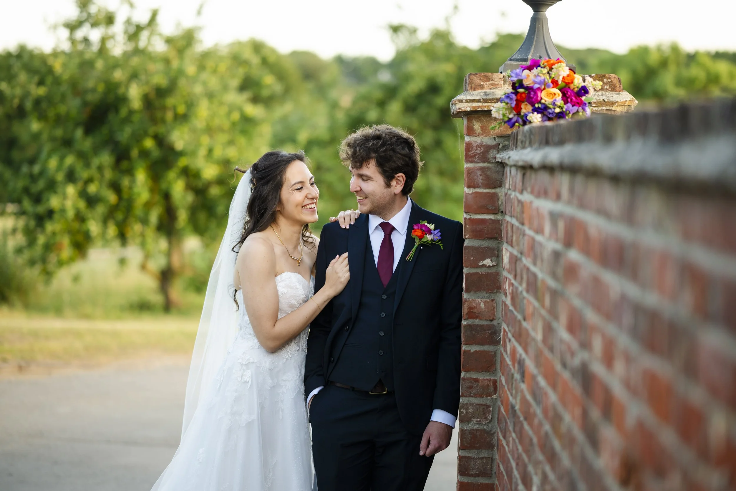 A bride and groom in wedding attire standing outdoors near a brick wall, smiling at each other, with a bouquet of colorful flowers on top of the wall.