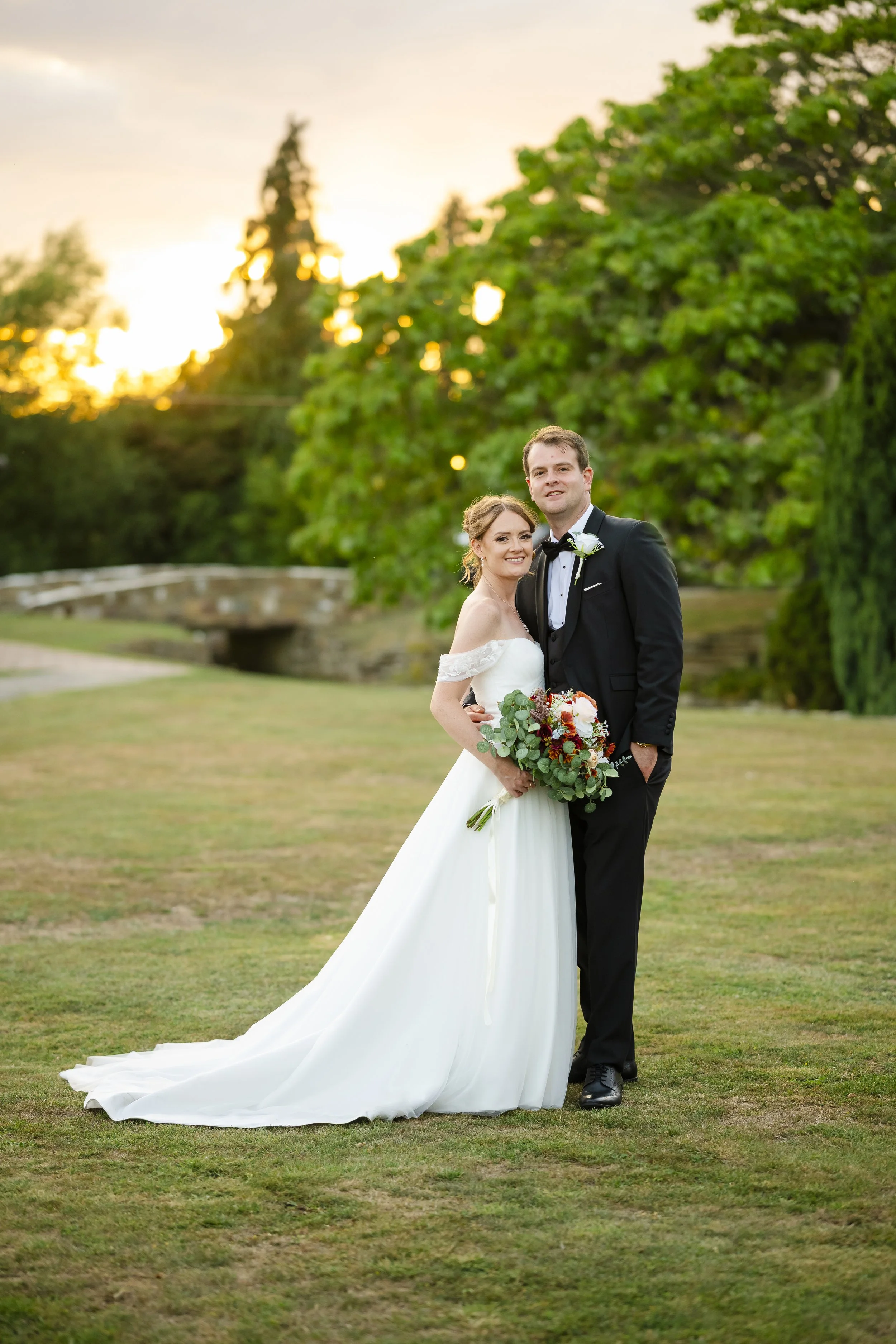A bride and groom stand together on a grassy area during sunset. The bride is in a white wedding gown holding a bouquet, and the groom is in a black tuxedo with a bow tie. They are smiling with trees and a stone bridge in the background.