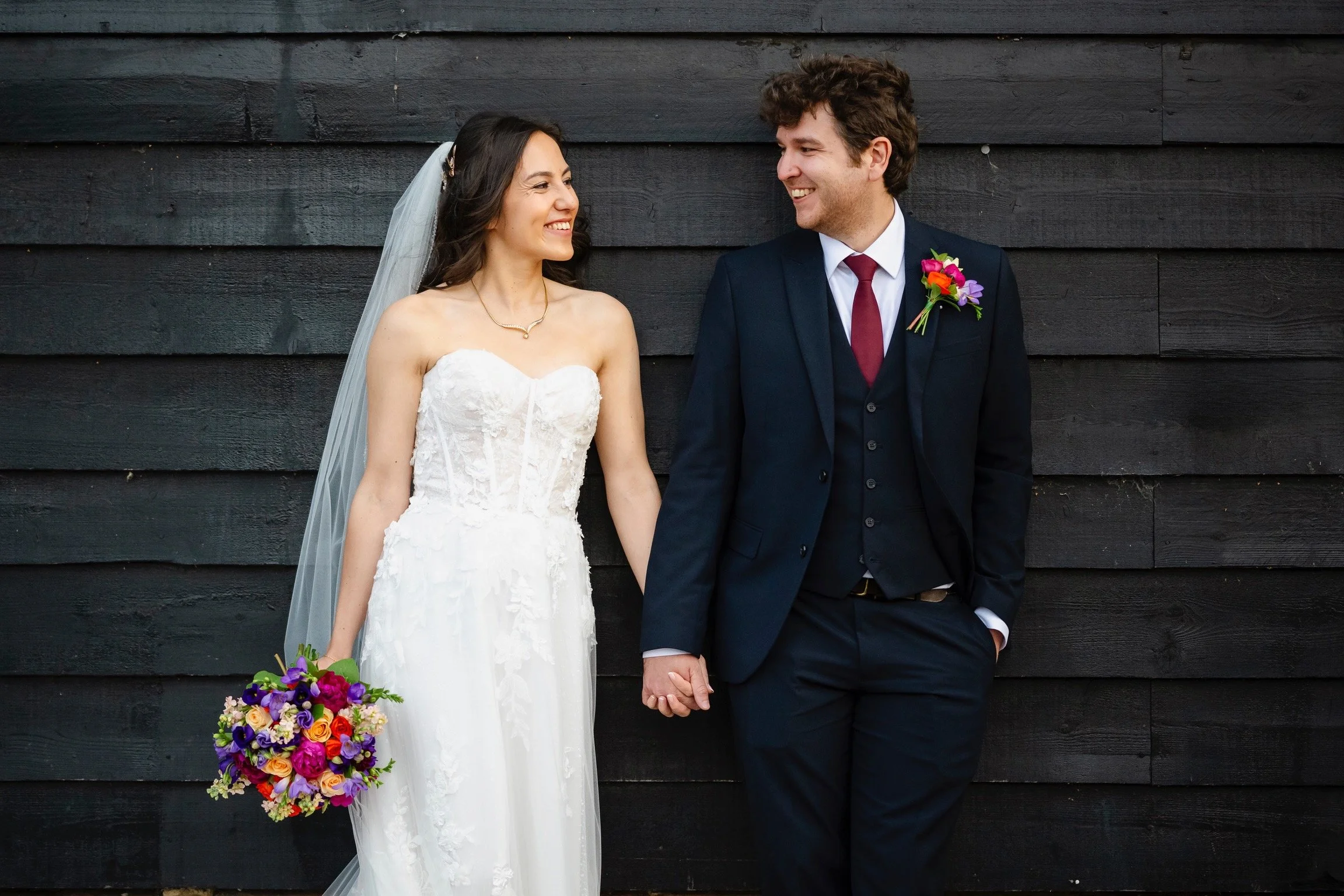 Bride and groom holding hands, standing against a black wooden wall, smiling at each other during a wedding.