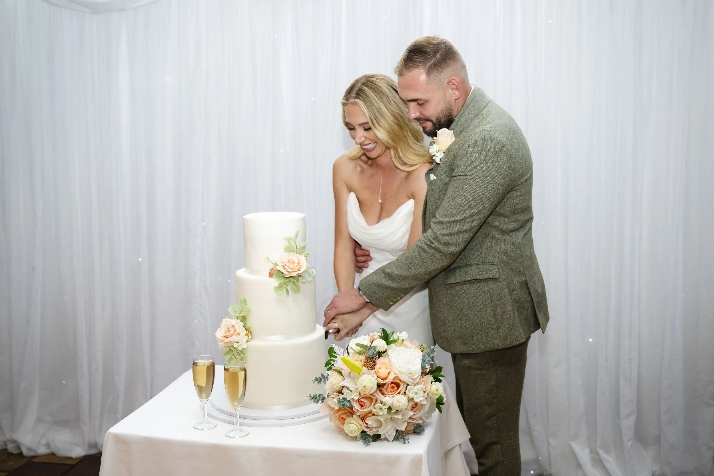 Bride and groom cutting wedding cake together at their reception, with floral bouquet and champagne glasses on table.