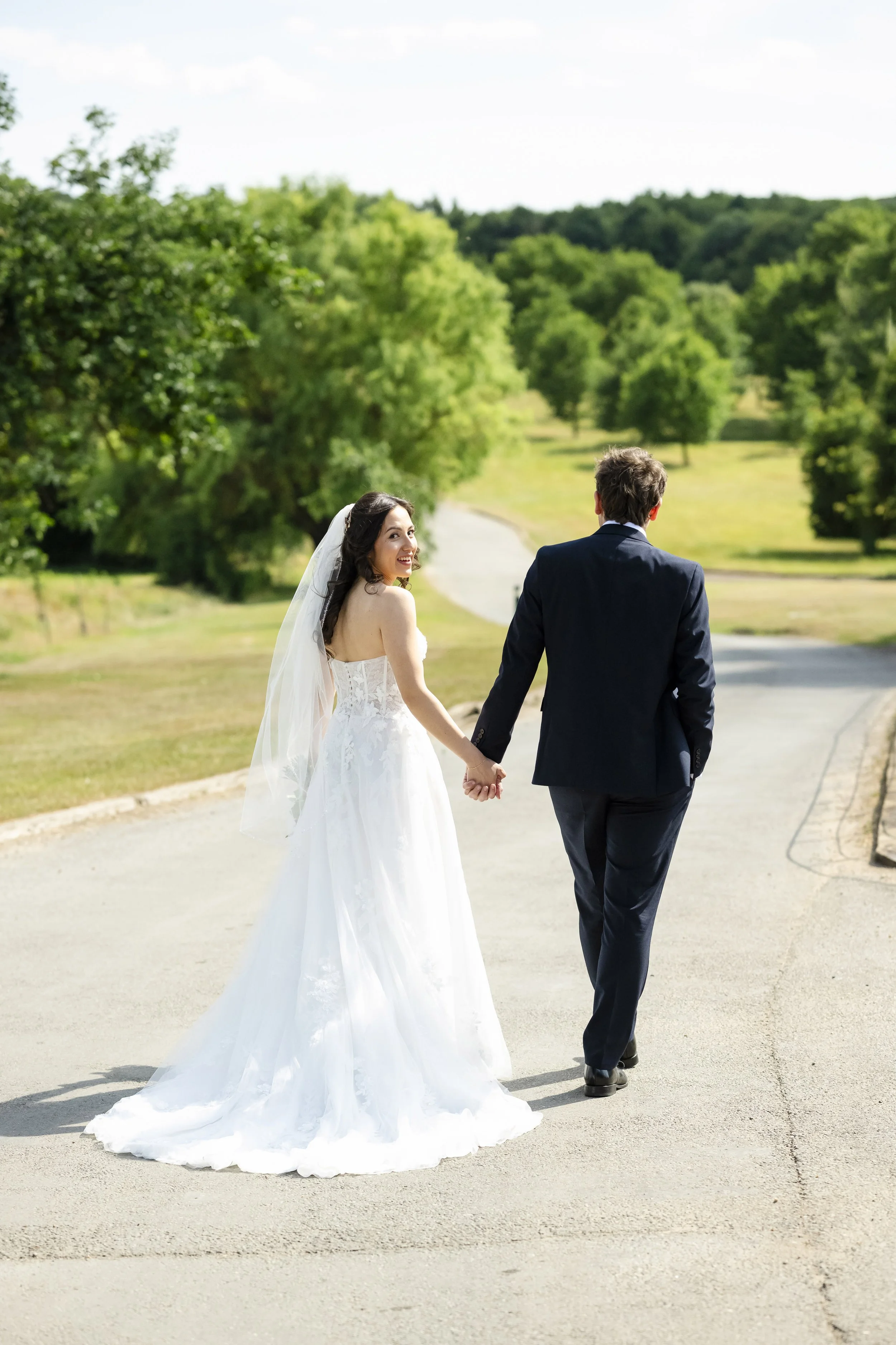 A bride and groom walking hand in hand on a paved path in a park or garden, surrounded by greenery and trees, during daytime.