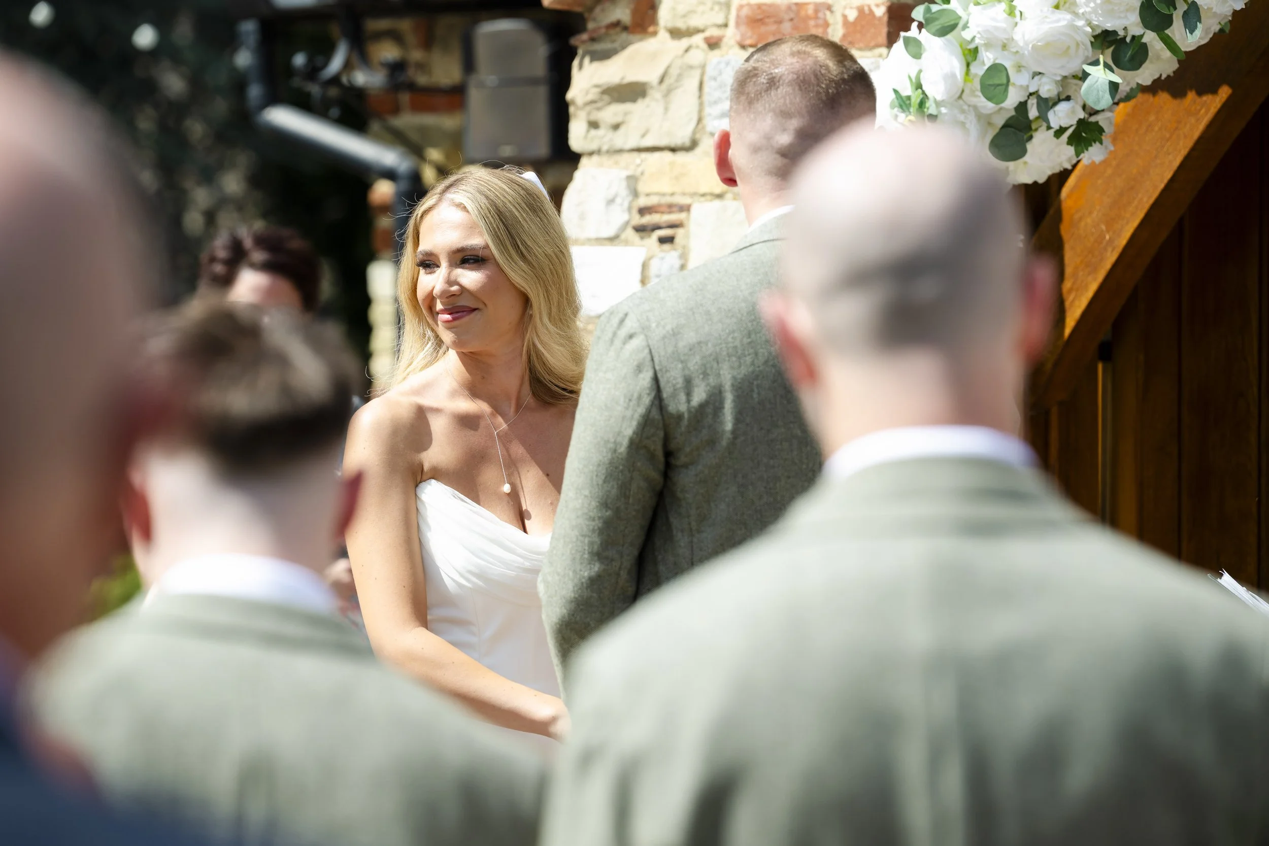 A woman dressed in white at a wedding ceremony outdoors, smiling as she looks at a man in a light gray suit, with several groomsmen in matching suits in the foreground. There are flowers and a rustic stone wall in the background.