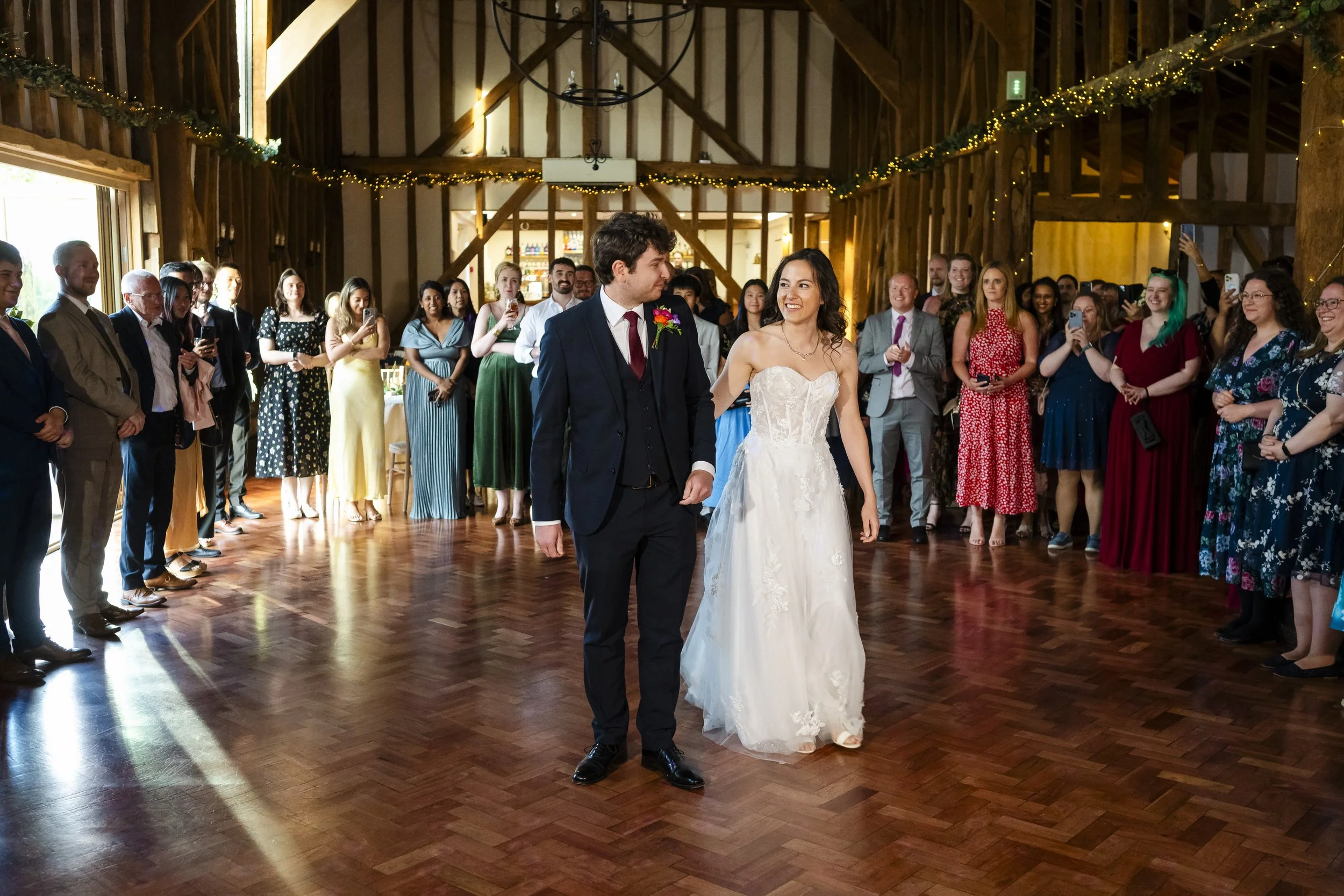 A newlywed couple is dancing together in a barn wedding venue with guests gathered around. The bride is wearing a white wedding dress and the groom is in a dark suit. The guests are standing in a circle, watching and some are taking photos, with fair