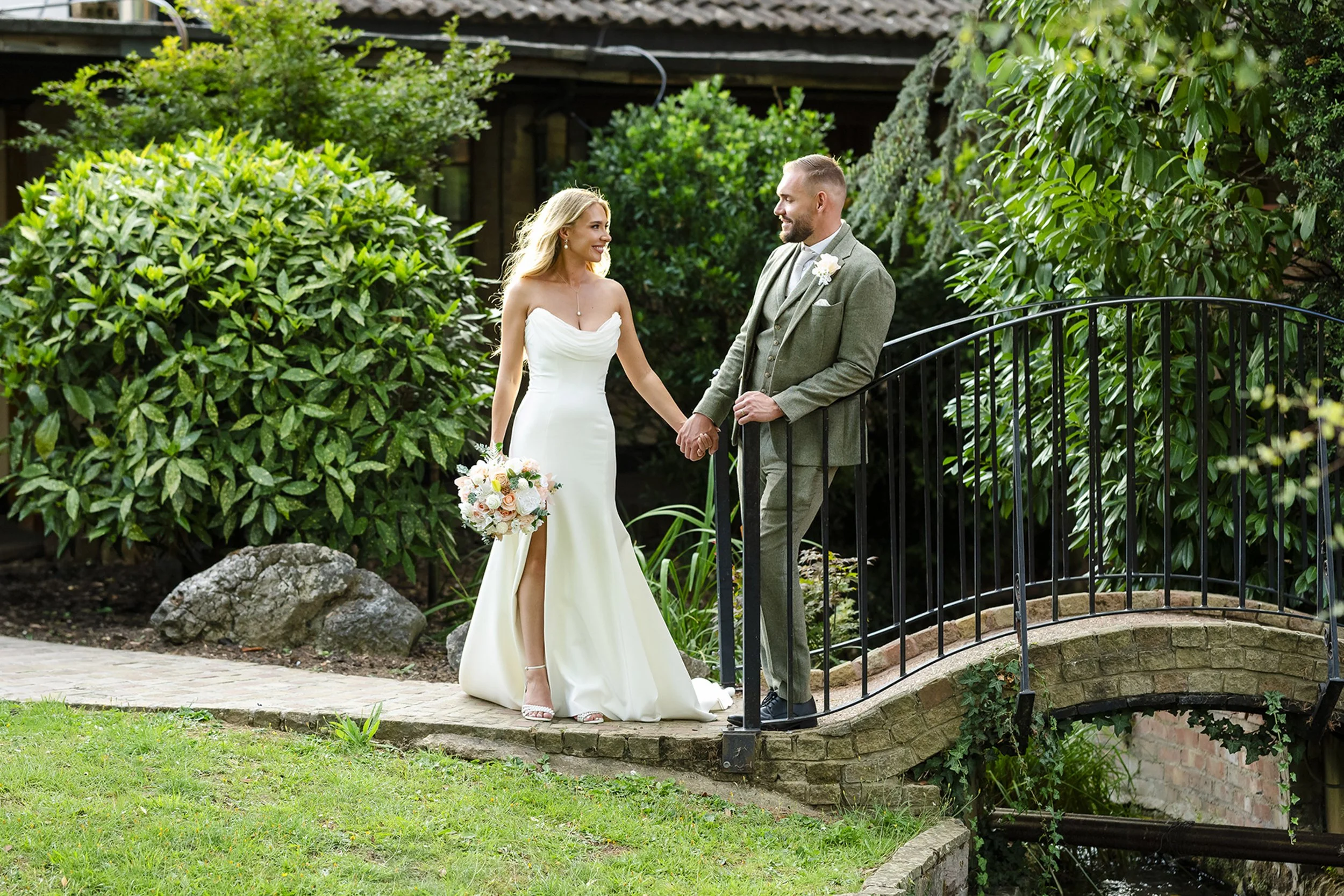 A bride and groom holding hands on a small stone bridge in a garden, surrounded by green bushes and trees, during their wedding photoshoot.