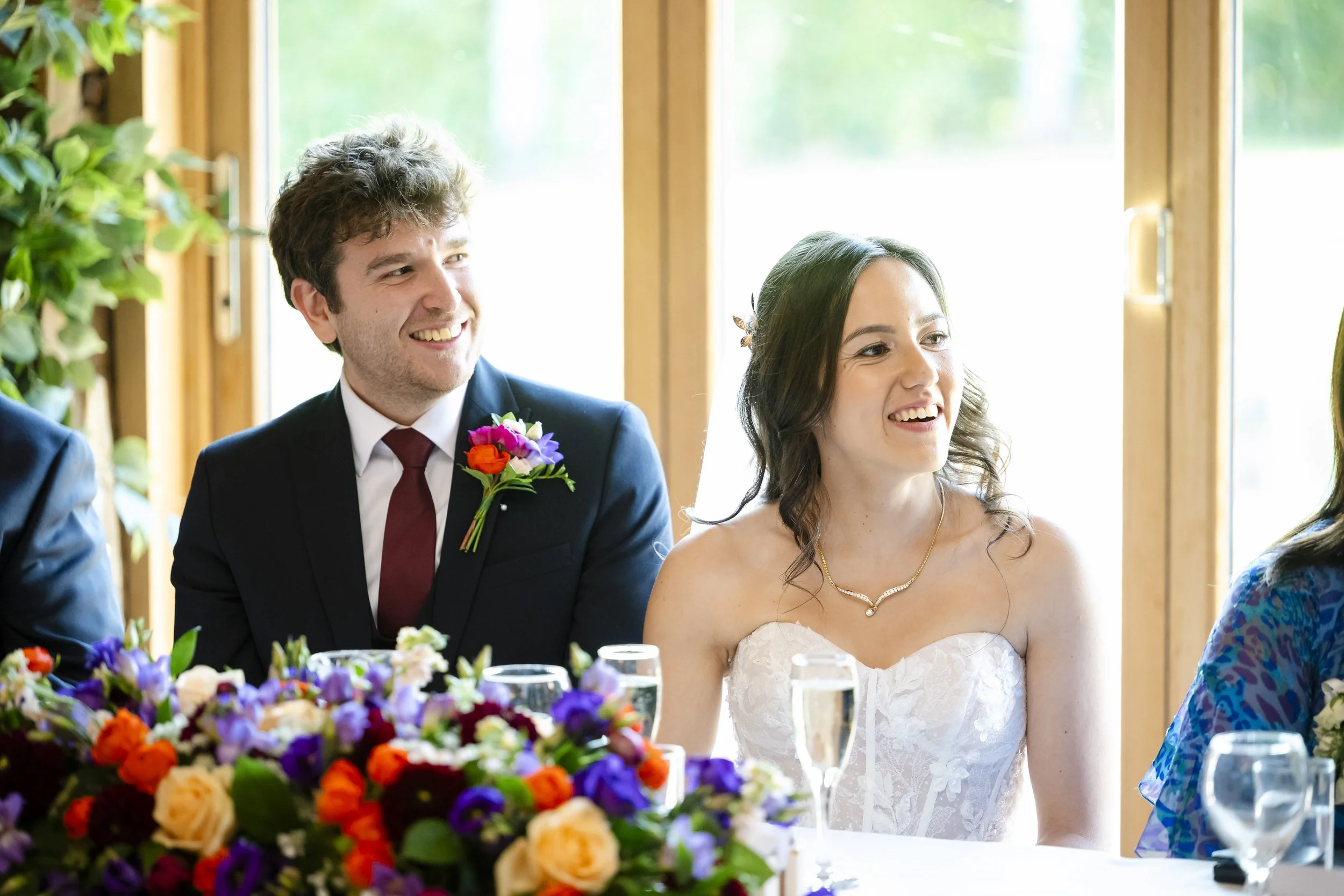 A bride and groom sitting at a wedding reception table, smiling, with a floral centerpiece and glasses of champagne in front of them.