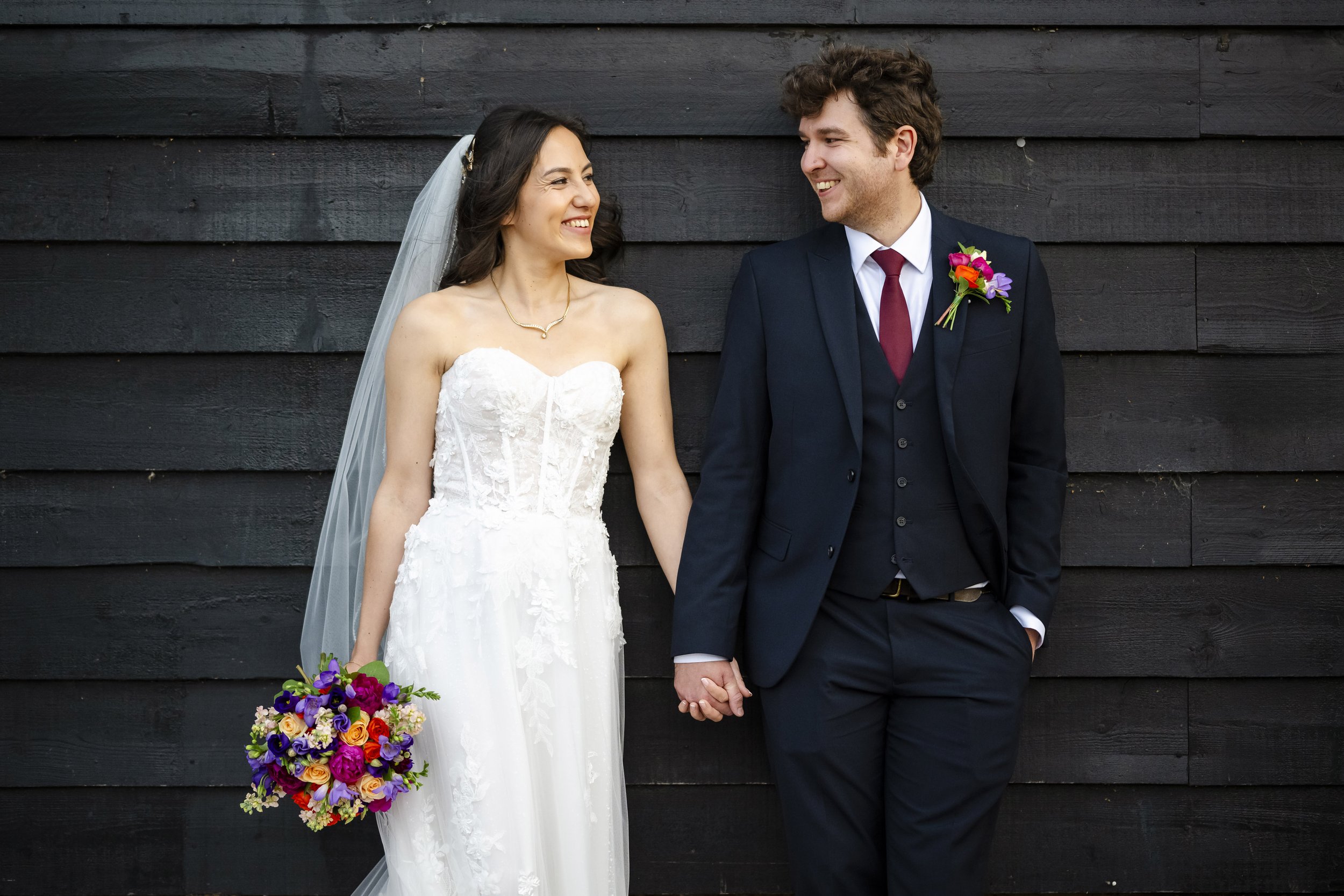 A bride and groom stand hand in hand, smiling at each other, against a dark wood wall. The bride wears a white lace strapless wedding dress, veil, and holds a colorful bouquet. The groom wears a navy suit with a red tie and a boutonniere with colorfu