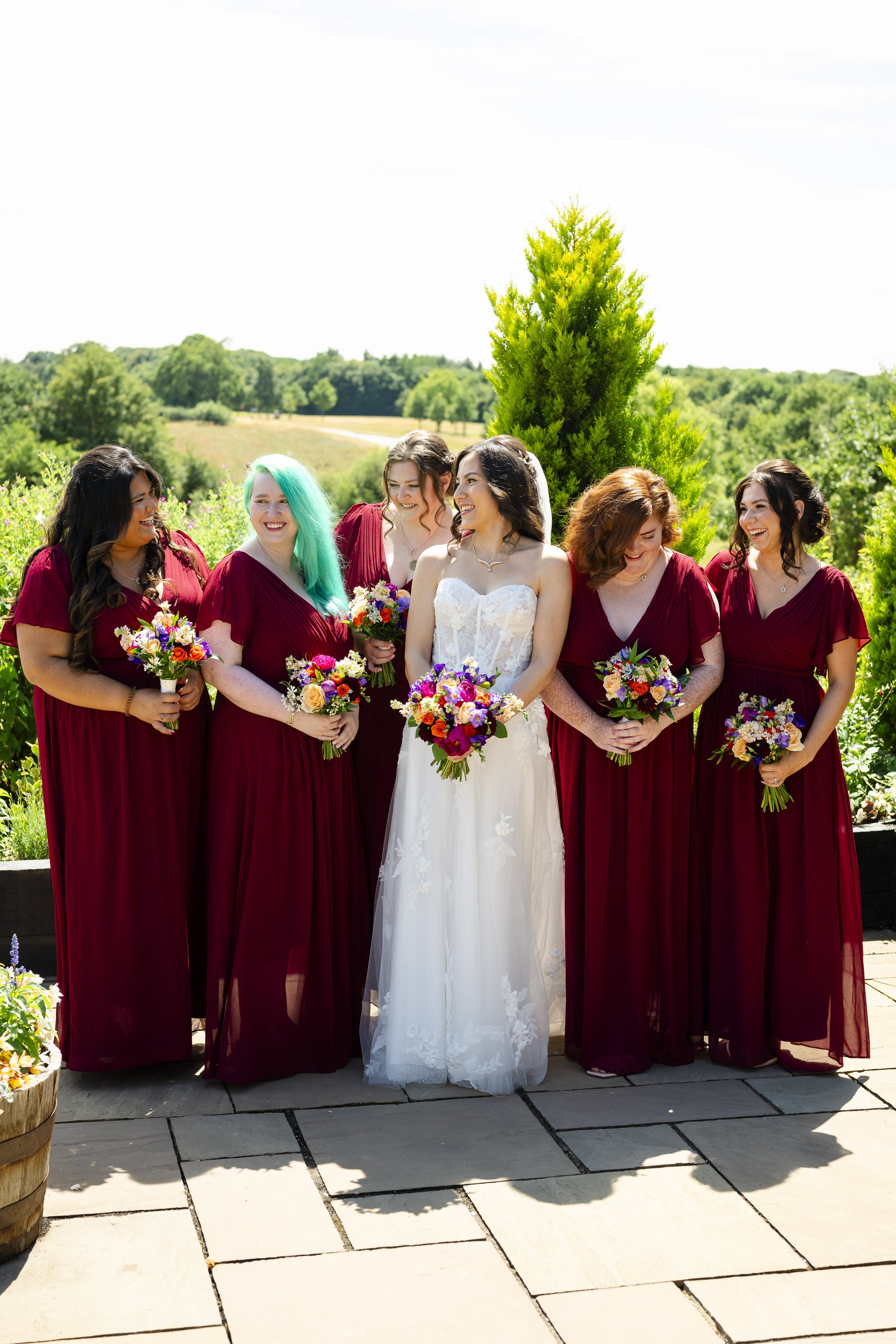 A bride in a white wedding dress and her bridesmaids in matching maroon dresses outdoors, holding bouquets of colorful flowers, smiling and laughing together.