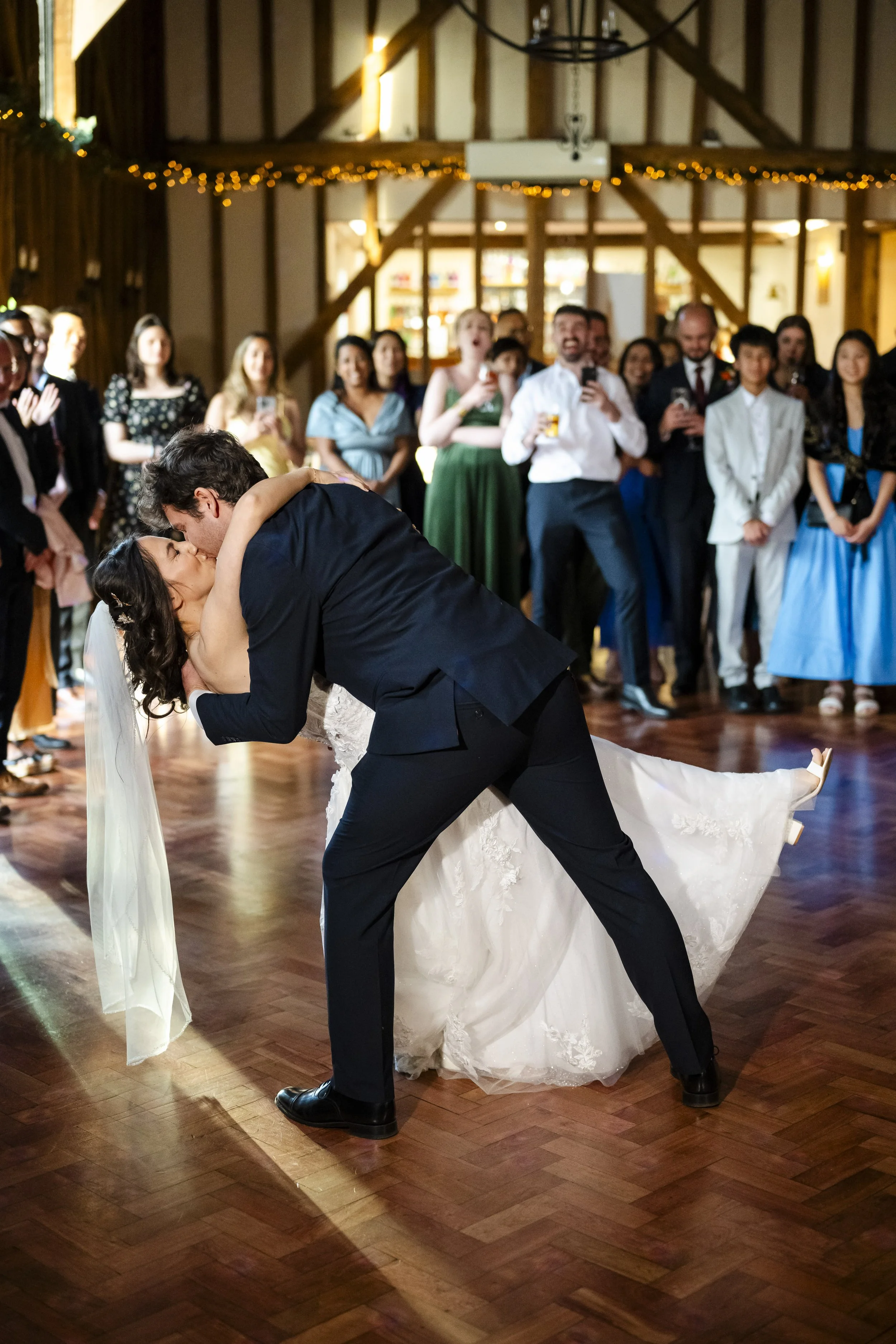 A bride and groom sharing a kiss during their wedding dance in front of guests in a rustic indoor venue decorated with string lights.