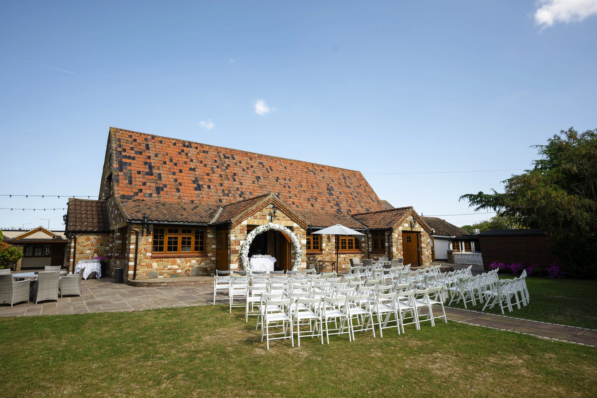 Outdoor wedding setup in front of a rustic brick building with a steep roof, decorated with white flowers and arranged chairs facing an arch, on a grassy and paved area under a clear blue sky.