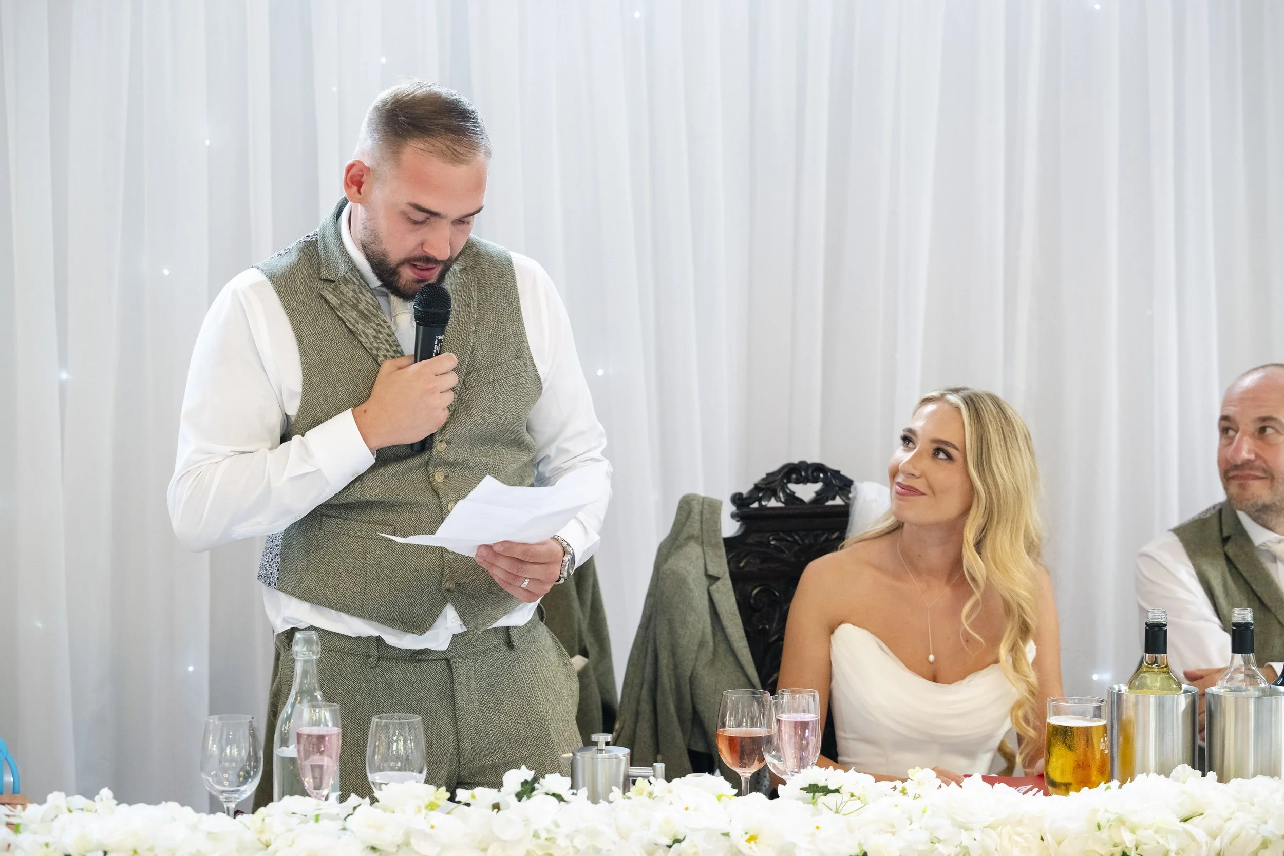 Man giving a speech with a microphone at a wedding reception, seated beside a woman in a white dress, with other guests and drinks visible on the table.