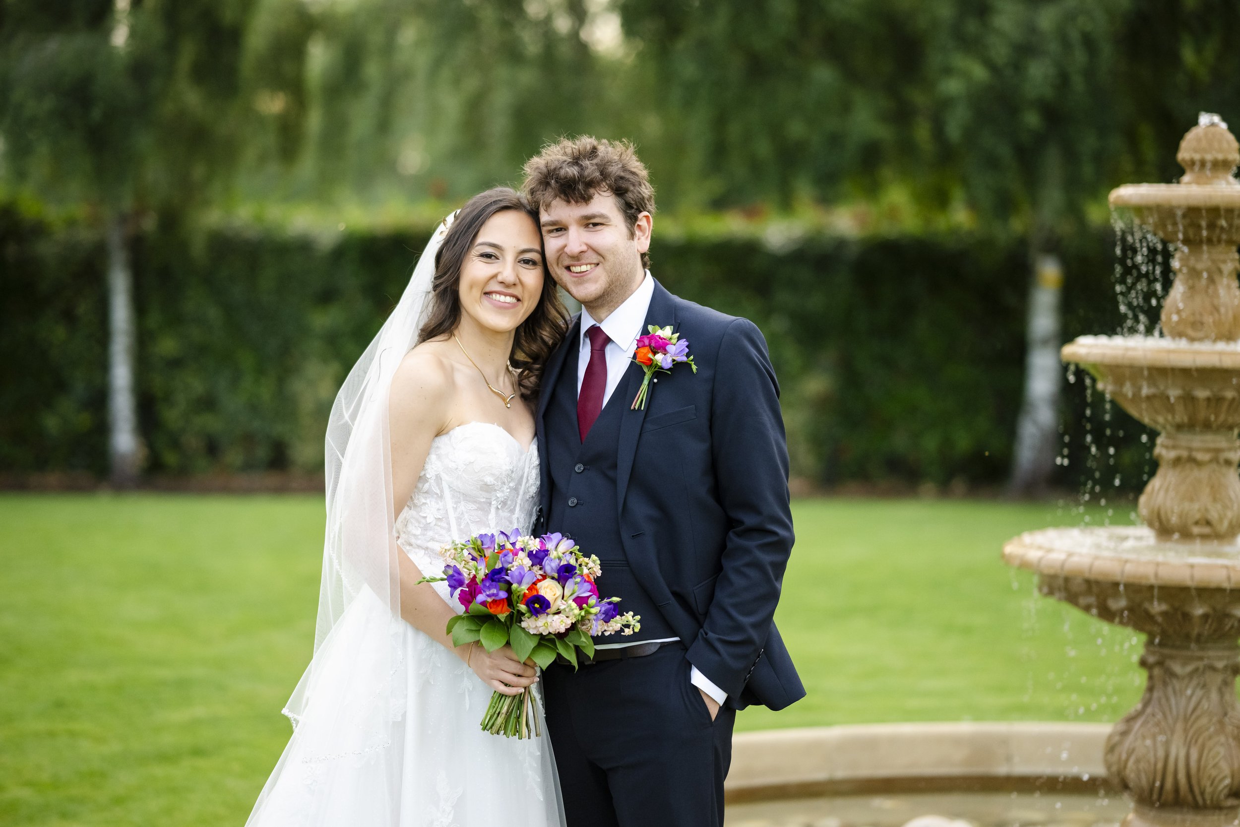 Smiling bride and groom standing outdoors near a fountain, with trees and greenery in the background.