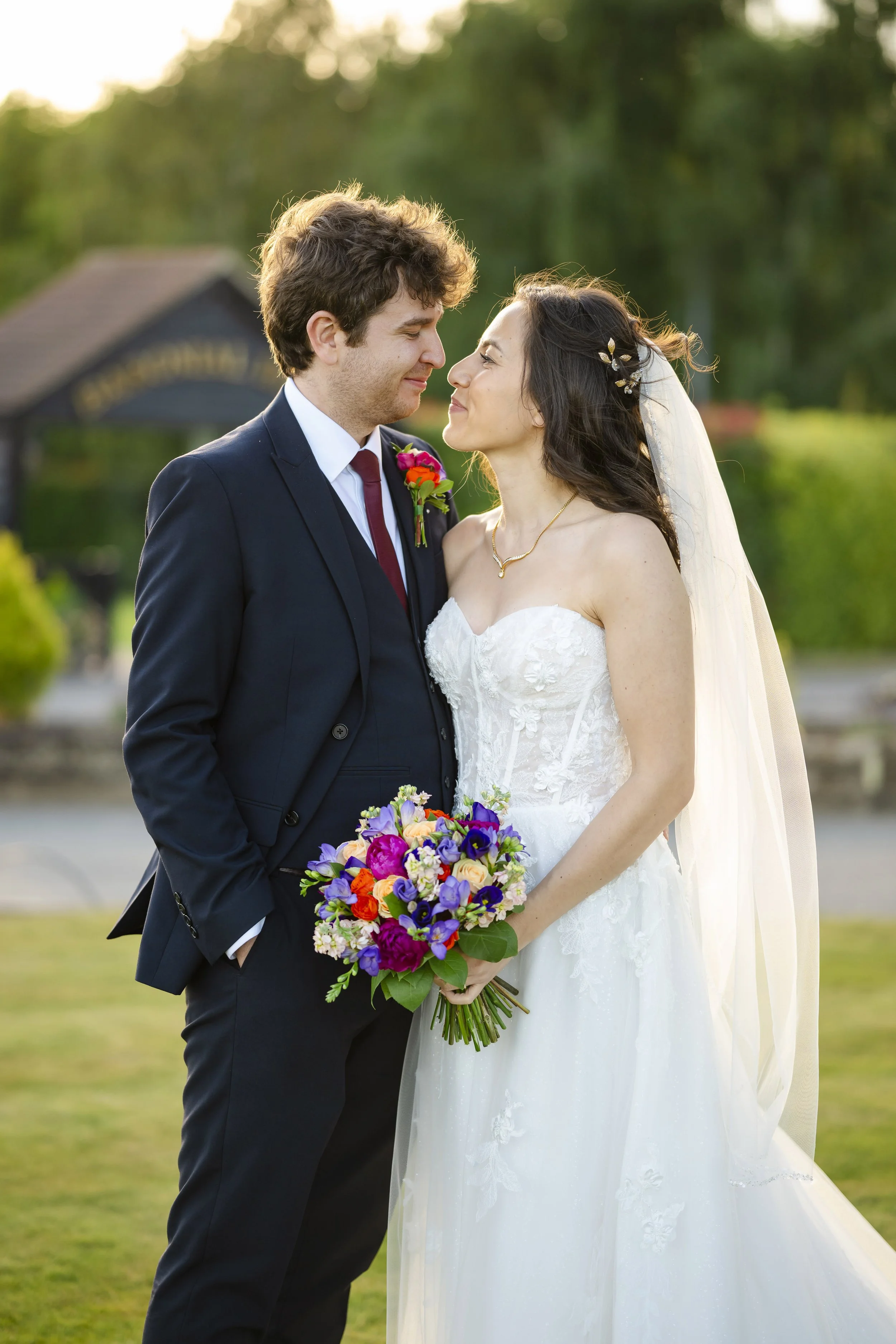A newlywed couple standing close together outdoors, gazing into each other's eyes, with a small building and trees in the background. The bride is holding a colorful bouquet of flowers.