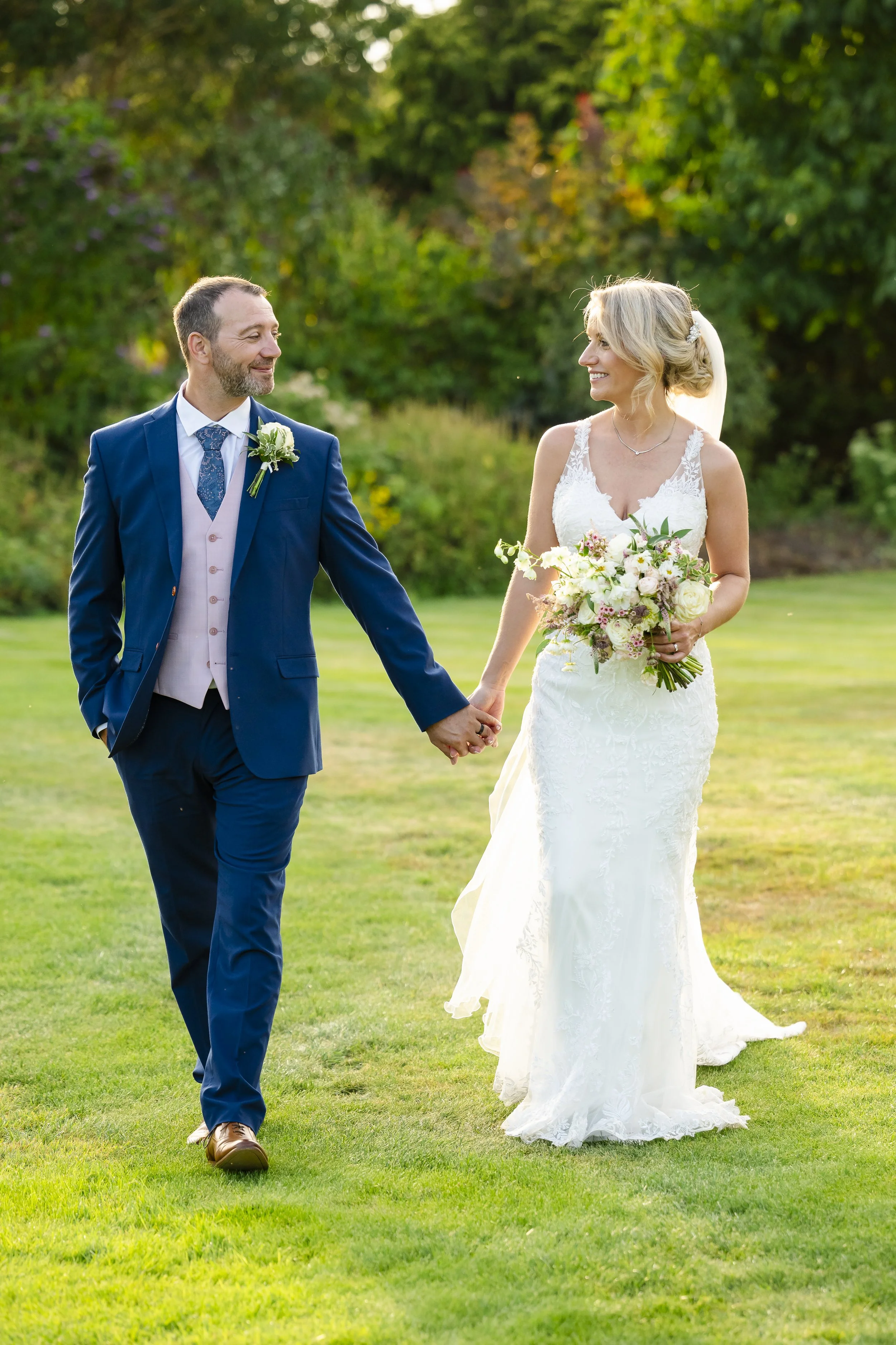 A newlywed couple walking hand in hand on a lush green lawn during their wedding day, with trees and colorful foliage in the background.
