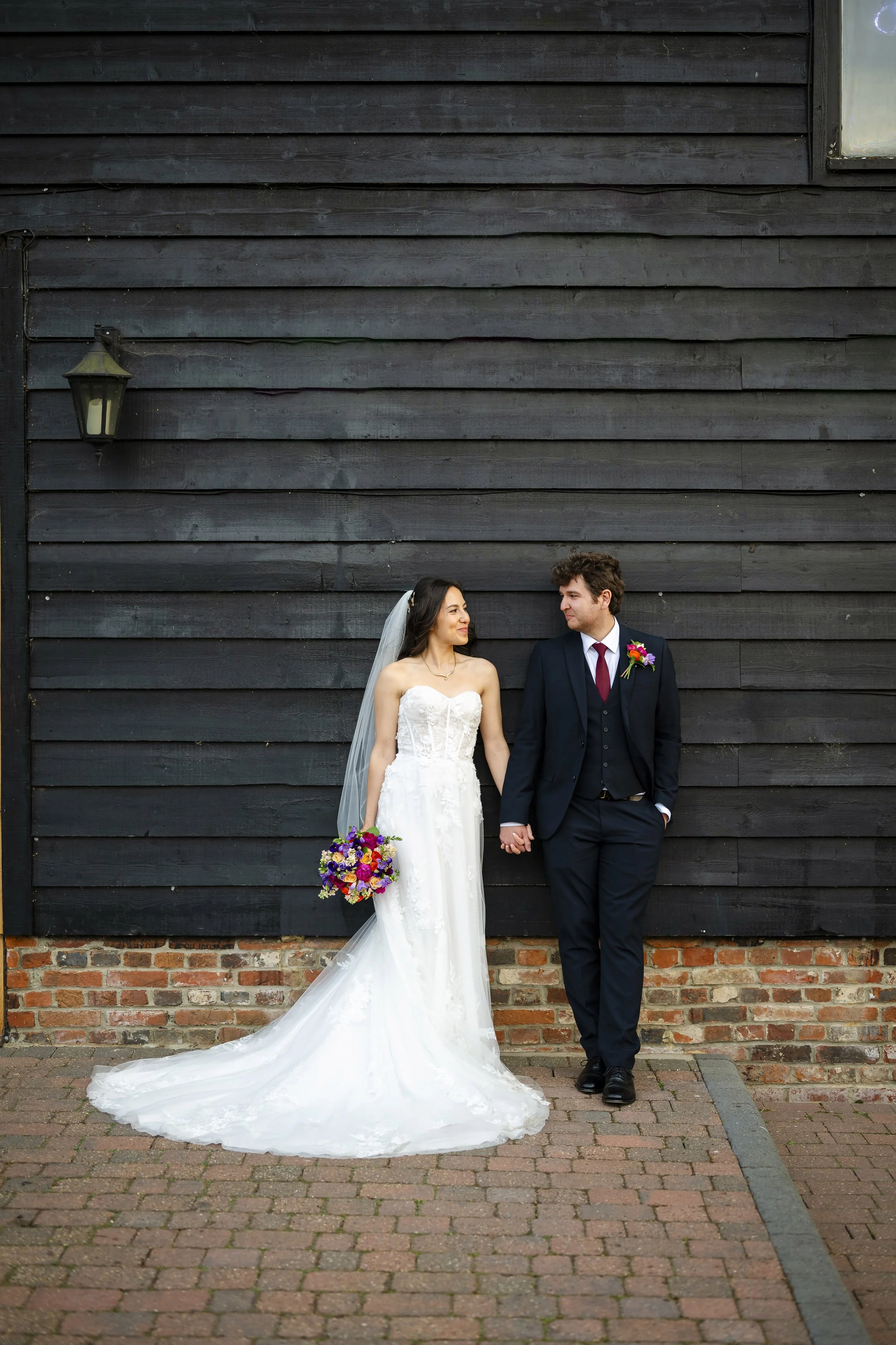 A bride and groom standing hand in hand, looking at each other, in front of a dark wooden wall. The bride wears a white strapless wedding gown with a long train and veil, while the groom is dressed in a dark suit with a red tie and a boutonniere. The
