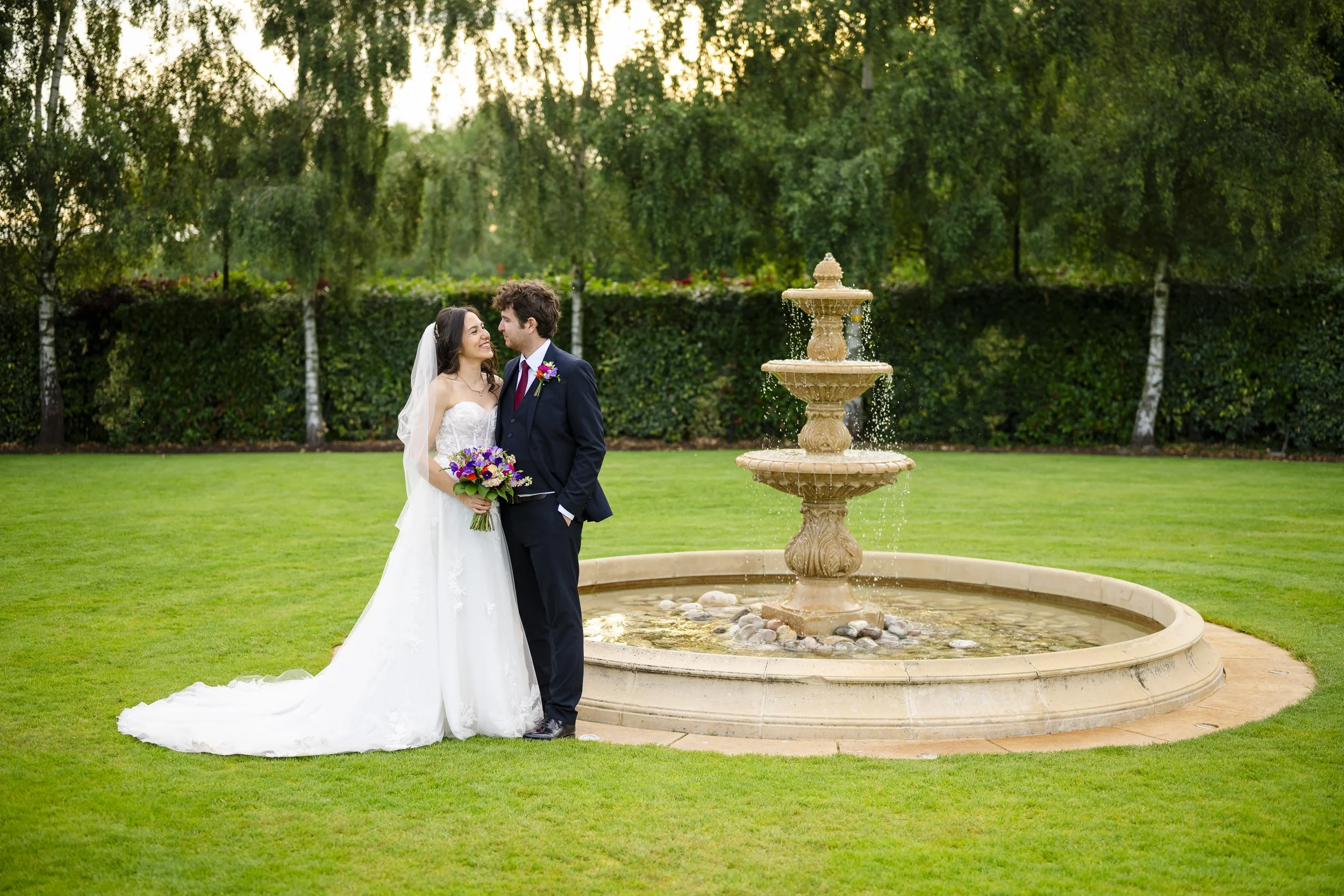 A bride and groom standing next to a fountain on a lush green lawn with trees in the background, holding a bouquet of flowers, during their wedding photoshoot.