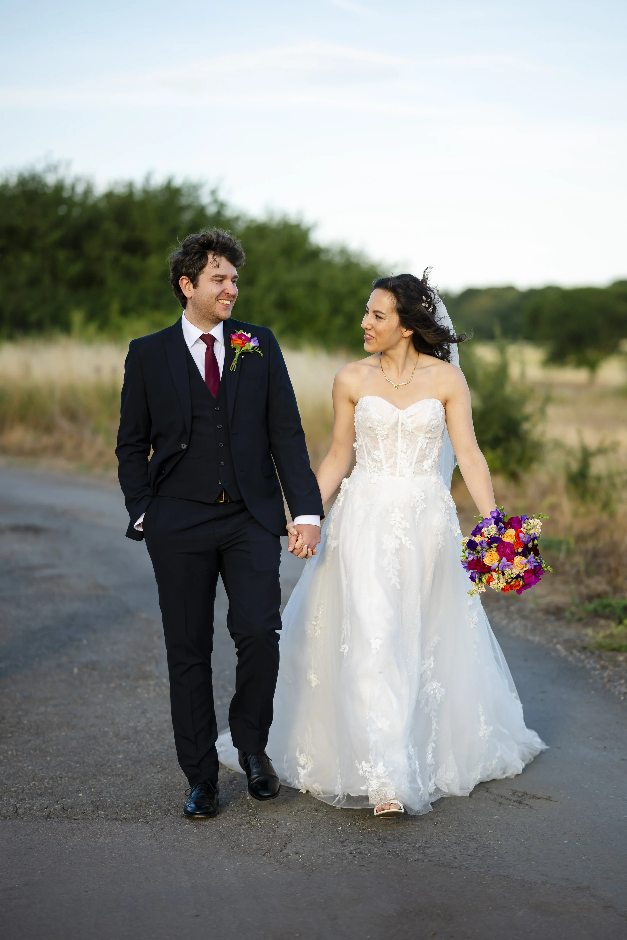 A bride and groom holding hands and walking outdoors, with the bride holding a colorful bouquet, on a rural road with trees in the background.