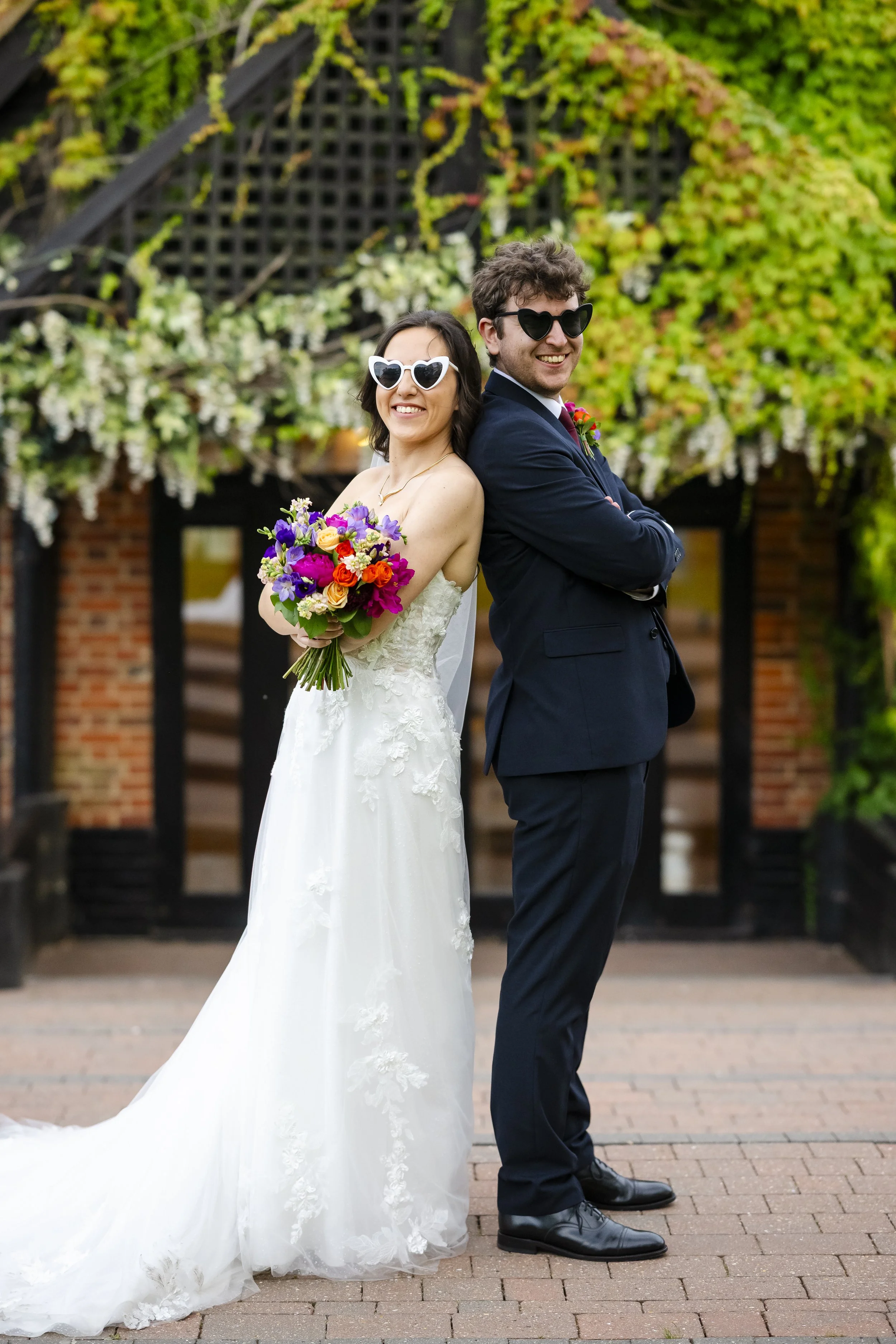 A bride and groom in wedding attire, standing back to back with arms crossed, wearing sunglasses and smiling, outdoors with greenery and a brick building in the background.