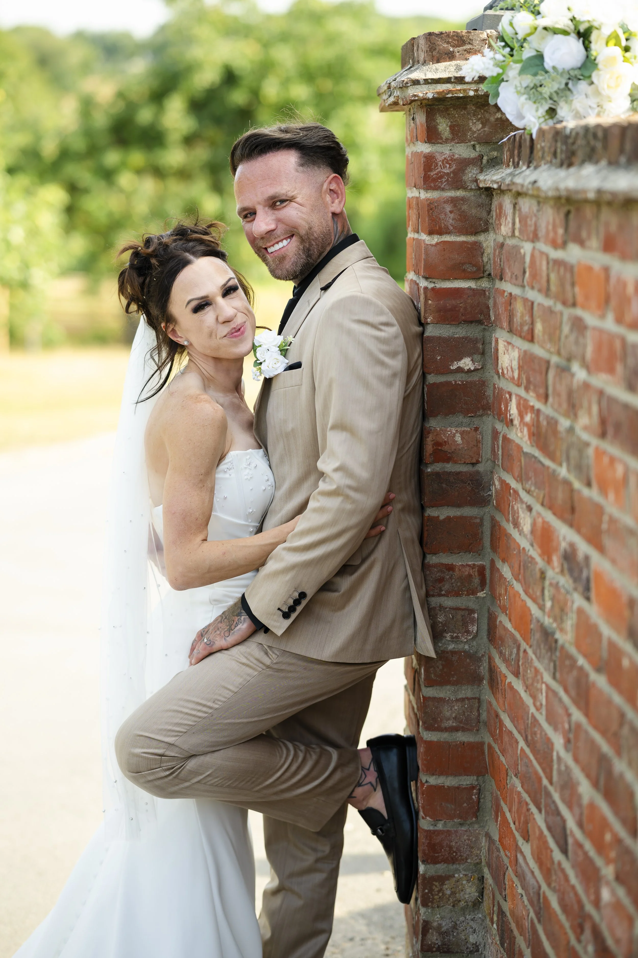 A happy bride and groom leaning against a brick wall outdoors on a sunny day.