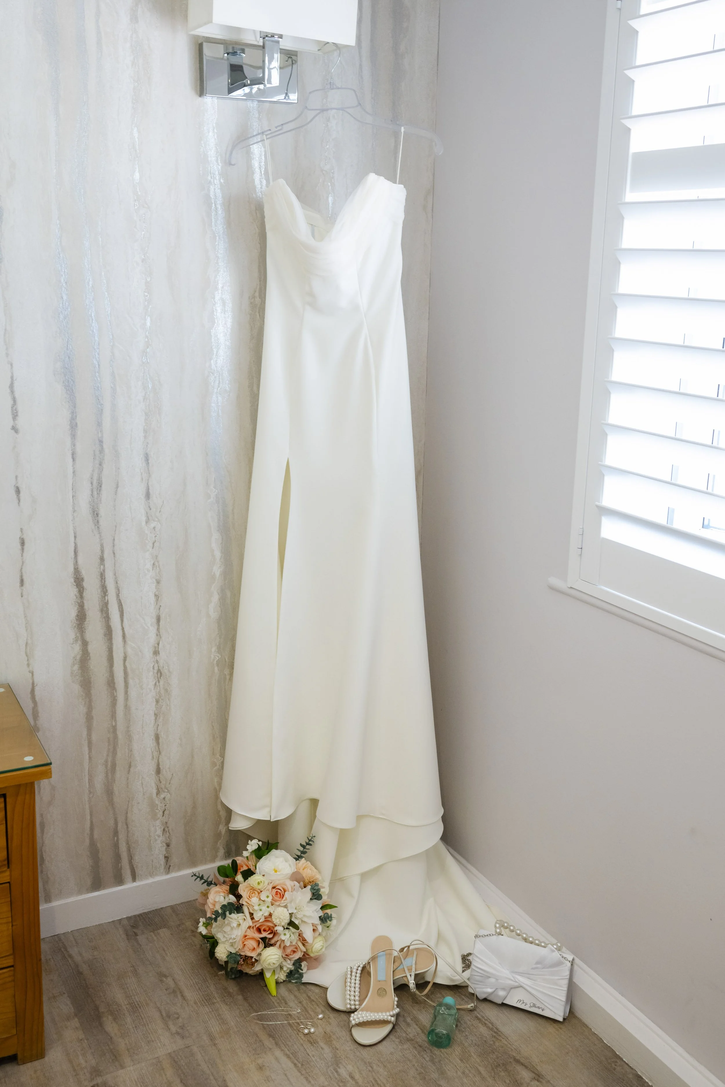 A white wedding dress hanging on a clear plastic hanger on the wall near a window with white shutters. A bouquet of pastel roses with greenery placed on the floor, along with a pair of white pearl-embellished shoes, a small white purse, a pearl neckl