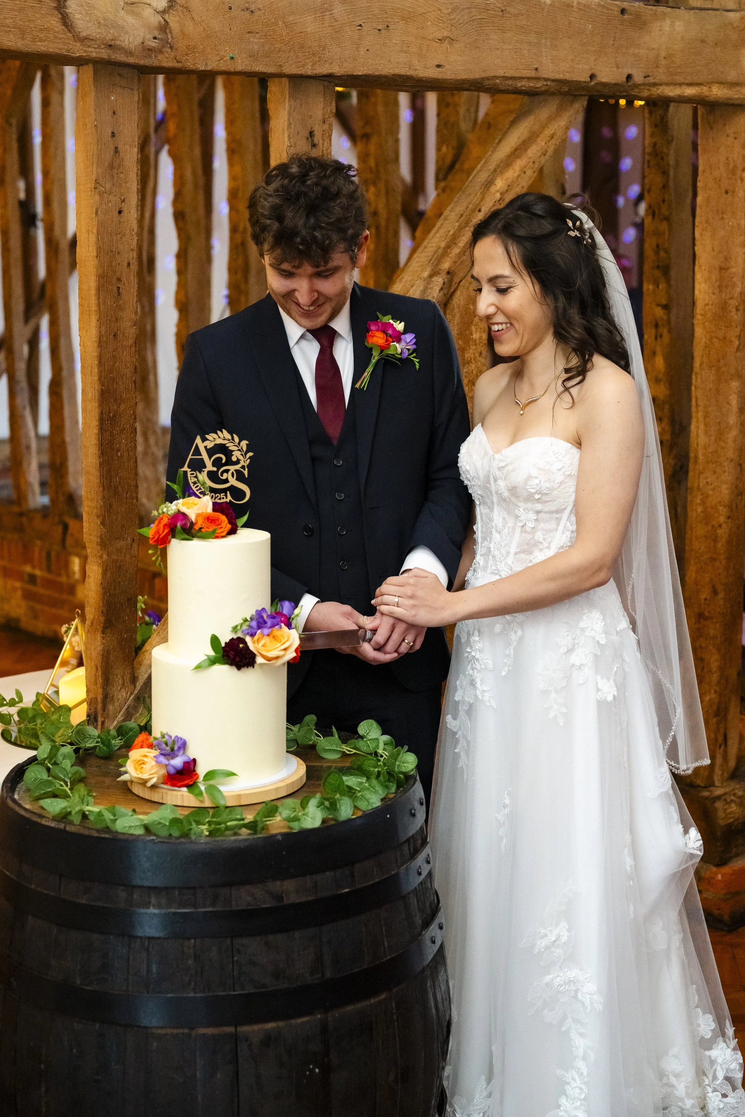 A bride and groom cutting a wedding cake together inside a rustic venue with wooden beams.